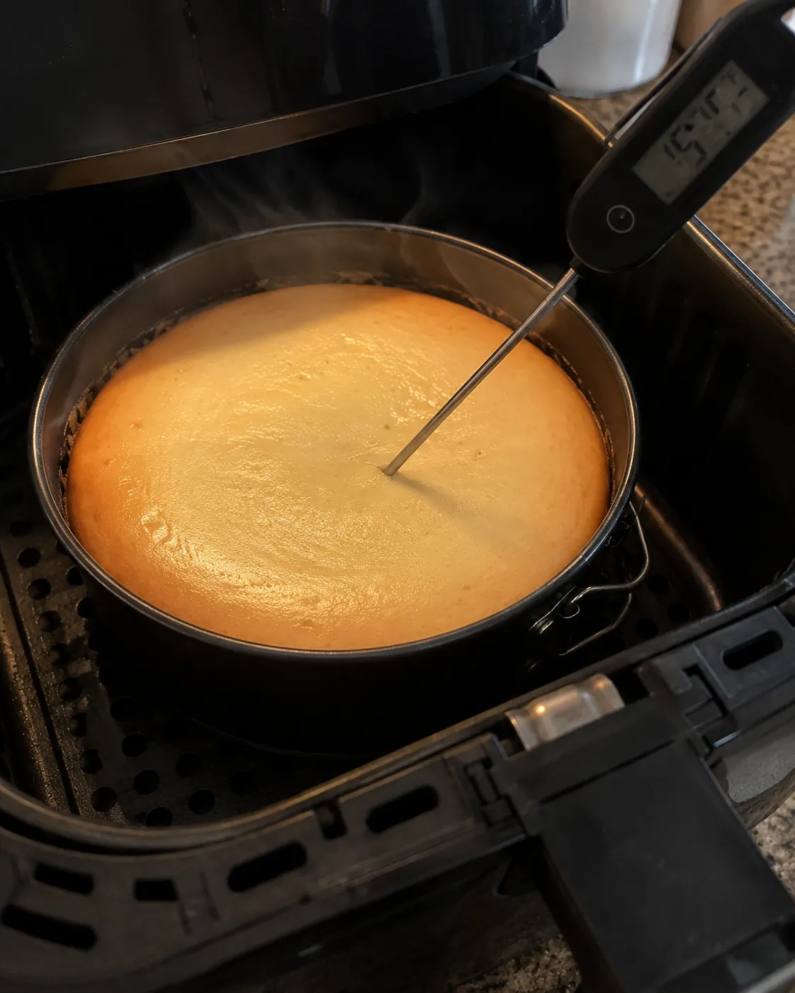 An instant-read thermometer probe pushed into the center of a cheesecake in a springform pan inside an air fryer basket