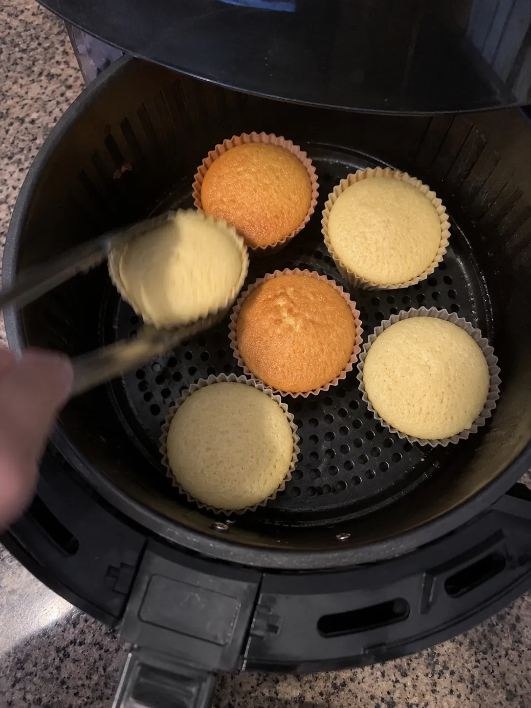Tongs lifting a pale cupcake out of an air fryer basket partway through the bake, with two deeply golden cupcakes and three pale ones still in place