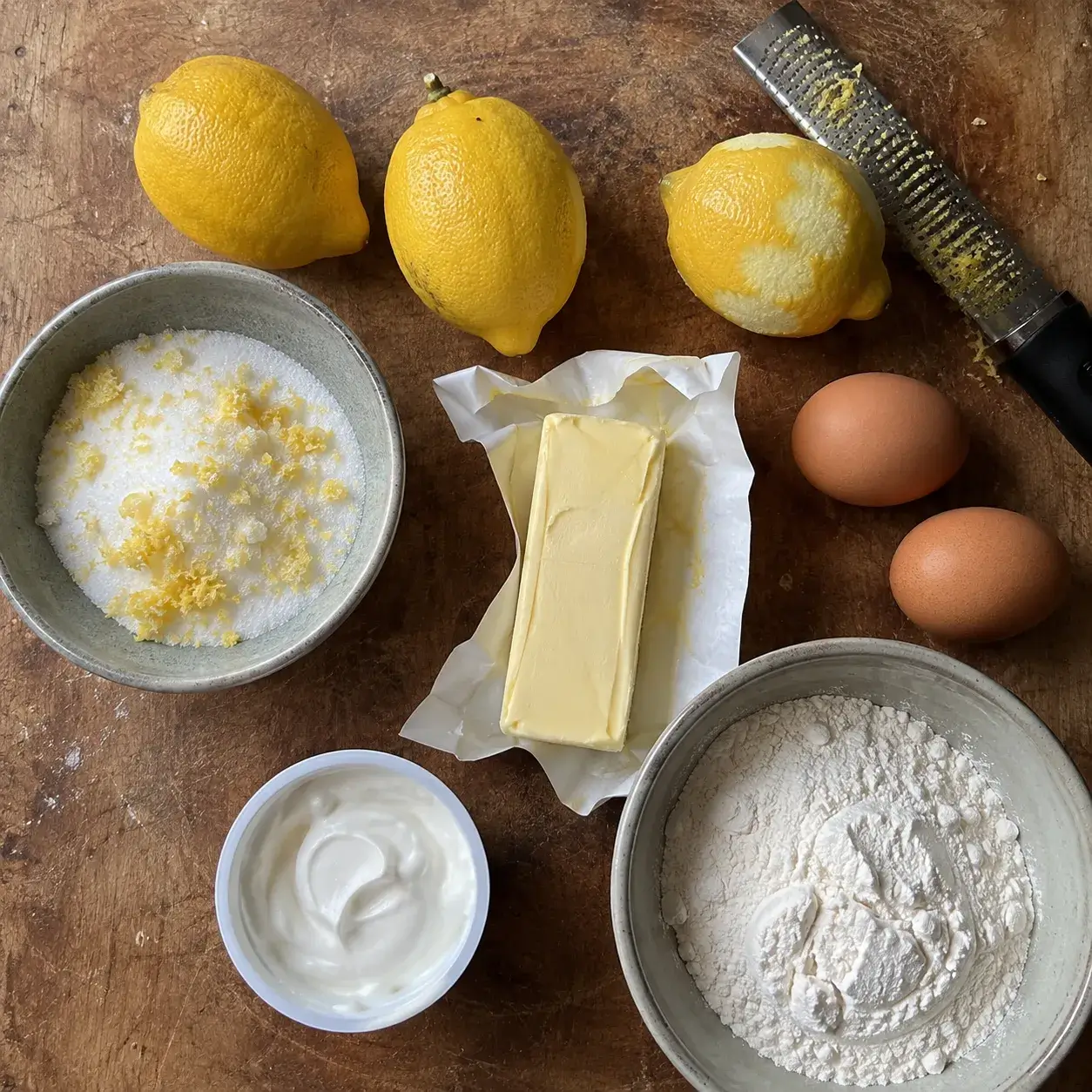 Ingredients for air fryer lemon cake laid out on a wooden counter, including three lemons, butter, eggs, flour and sour cream, with one lemon already zested next to a bowl of zested sugar