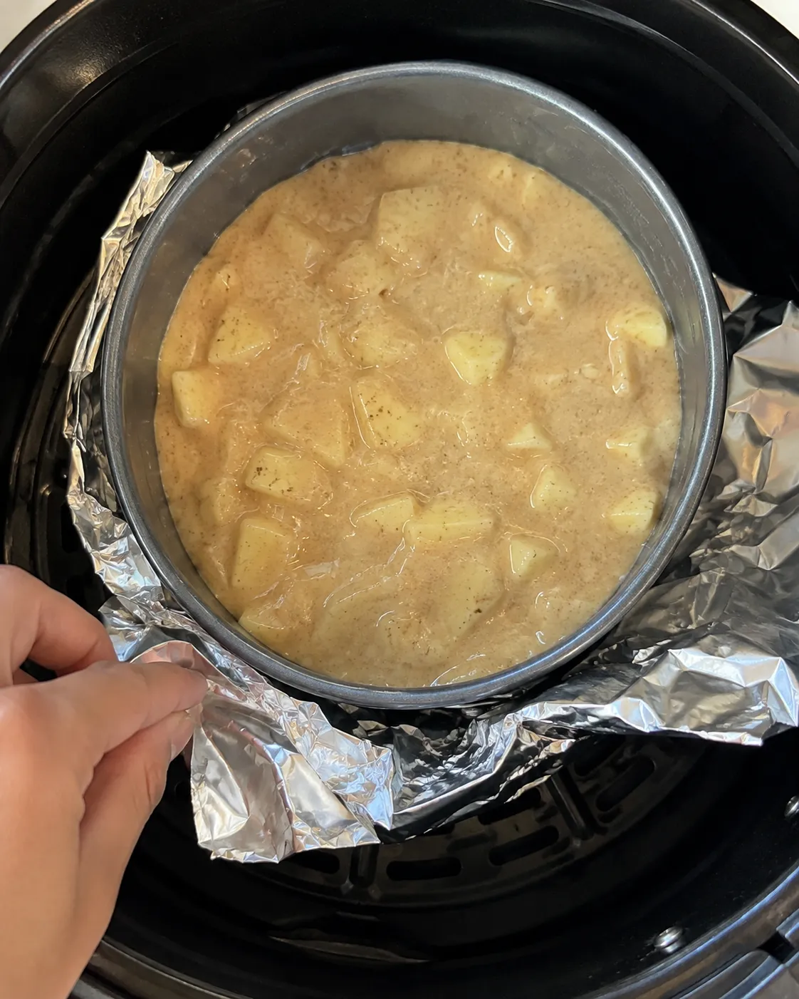 A tin of apple cake batter sitting in an air fryer basket with foil folded around the base of the tin