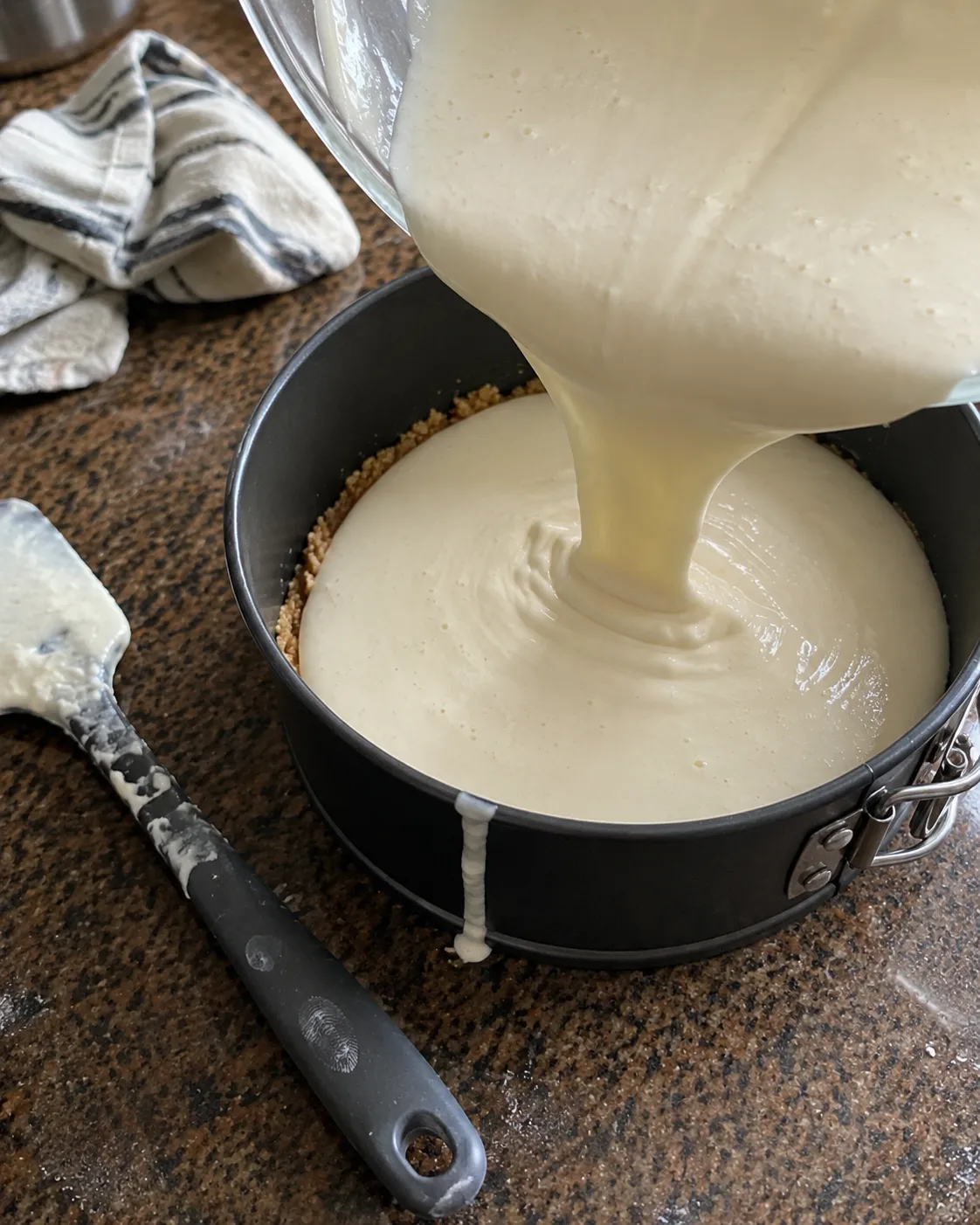 Smooth cheesecake batter being poured from a glass bowl onto a baked crumb crust in a springform pan
