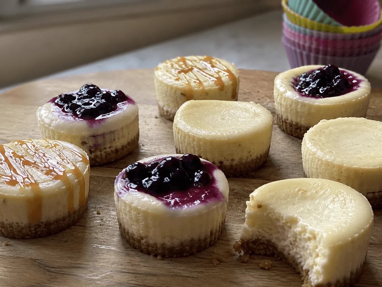 Air fryer mini cheesecakes on a wooden board topped with blueberry compote and caramel, one with a bite taken