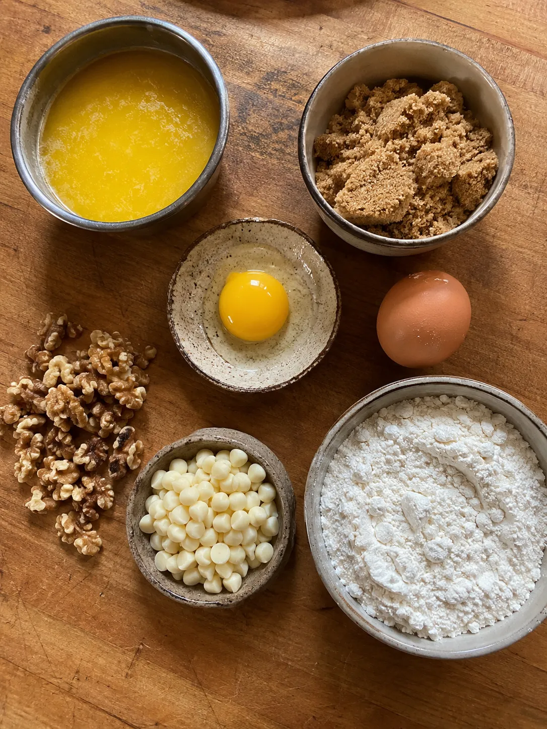Blondie ingredients in bowls: melted butter, brown sugar, an egg yolk, a whole egg, walnuts, white chocolate chips and flour