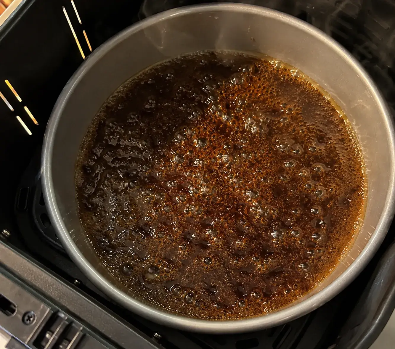Butter and dark brown sugar bubbling in a cake pan inside an air fryer basket, the syrup gone deep amber