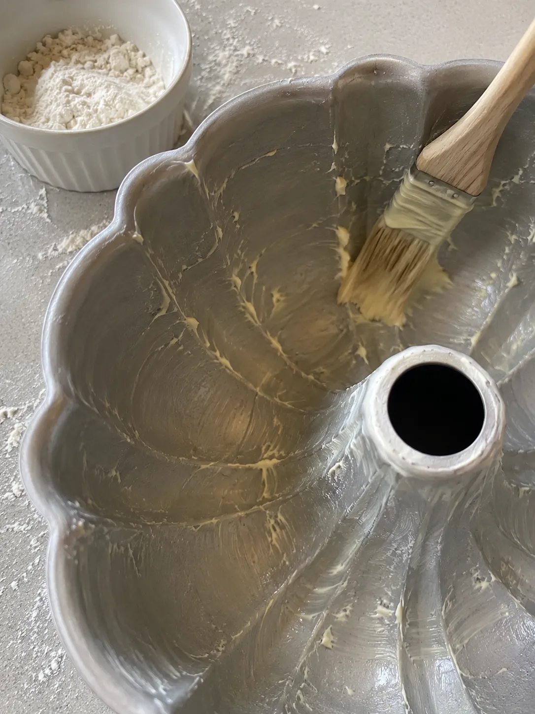 A pastry brush painting soft butter into every flute of a bundt pan, with a bowl of flour beside it and flour dusted on the counter