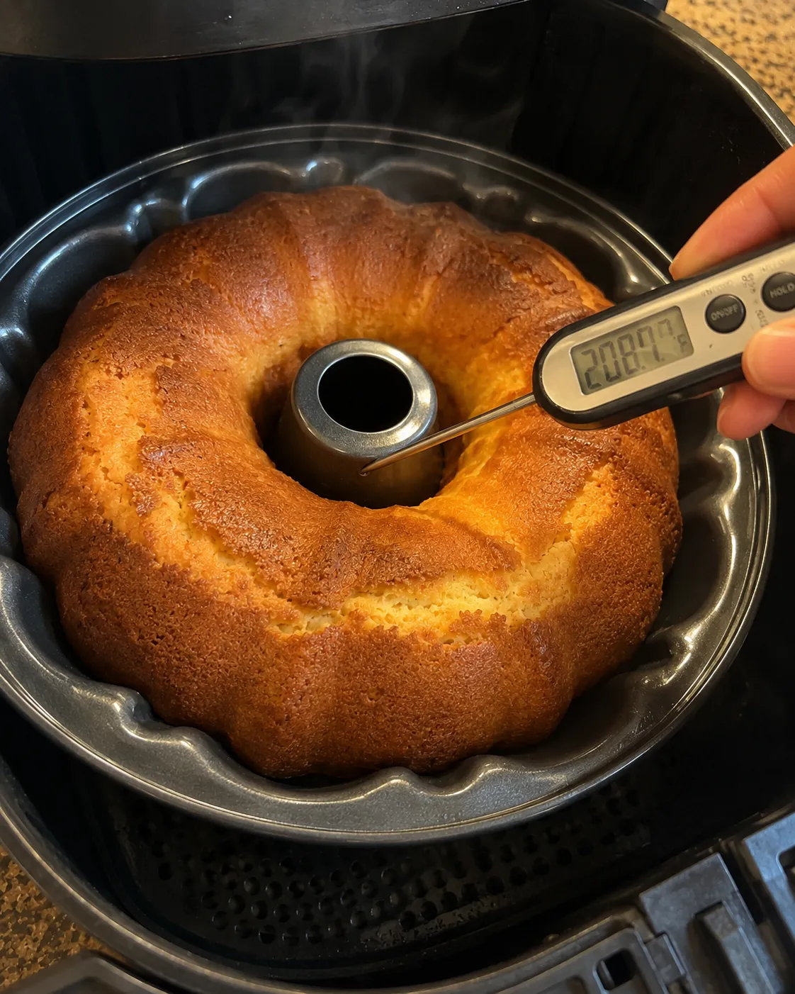 An instant-read thermometer probing a baked ring pound cake in an air fryer basket, the display reading 208.7 degrees Fahrenheit