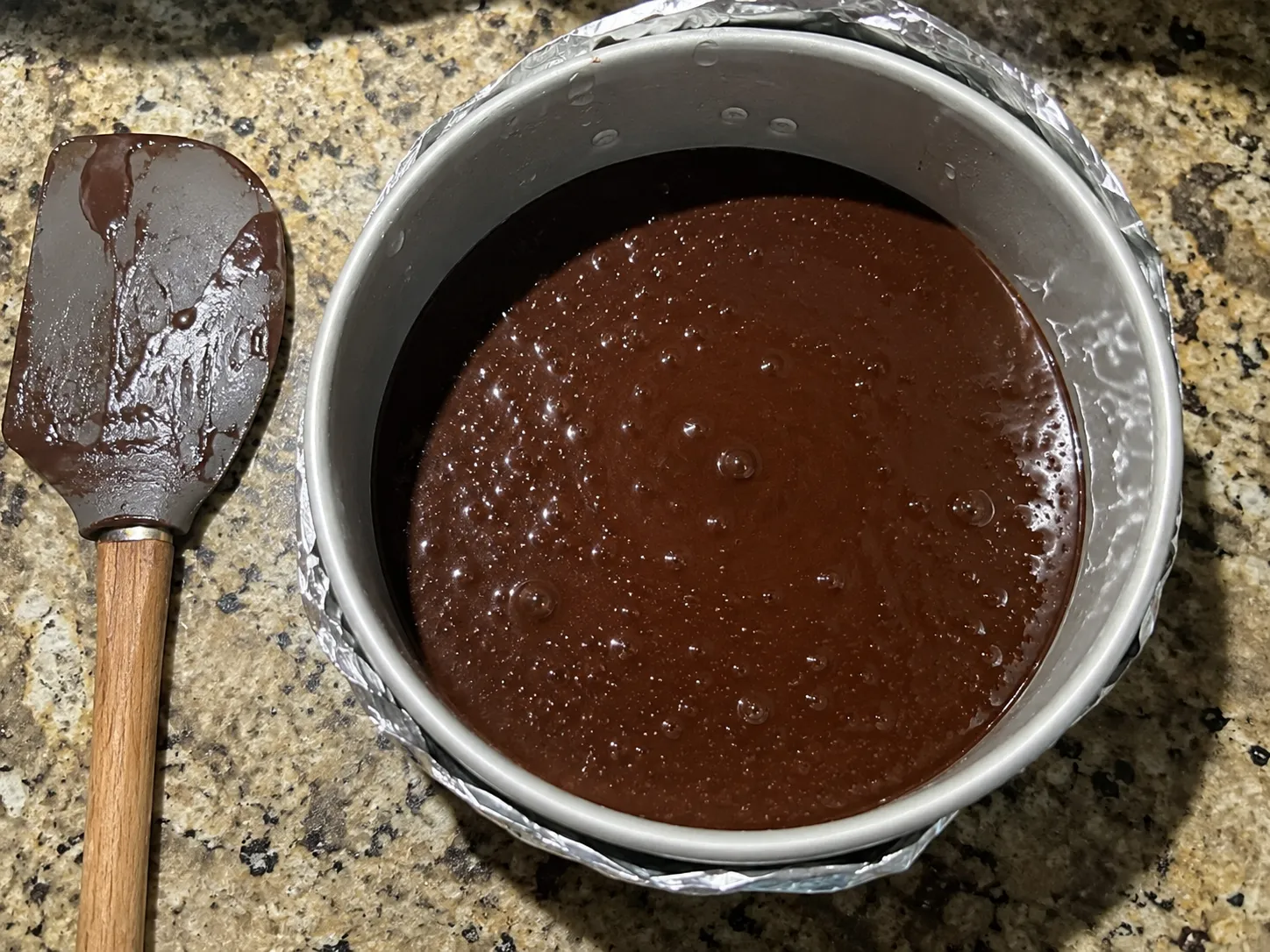 Overhead view of thin glossy chocolate batter poured into a round metal pan wrapped on the outside with a foil cake strip