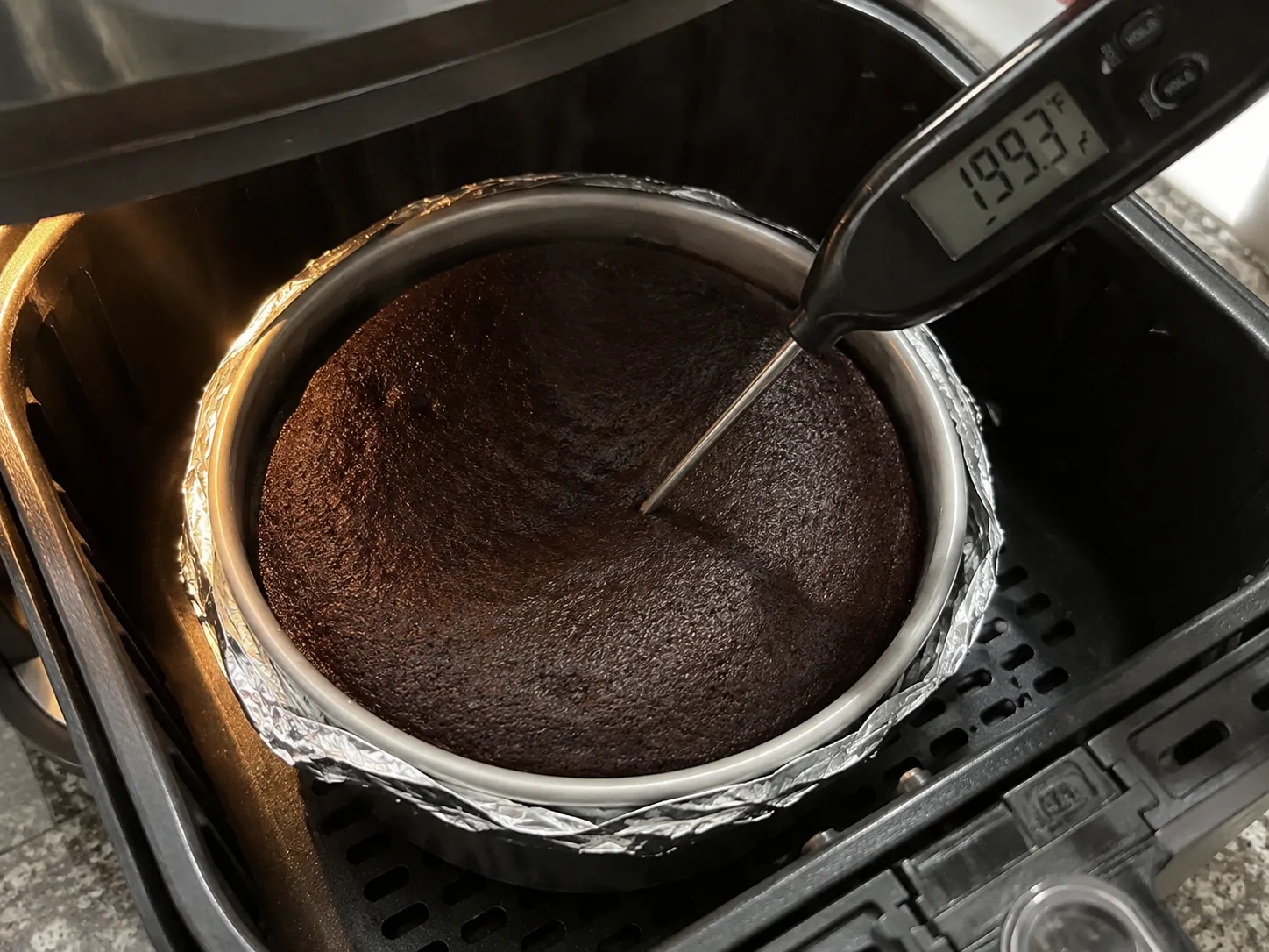 An instant-read thermometer pushed into the center of a baked chocolate cake in an air fryer basket, the display reading 199.3 degrees Fahrenheit