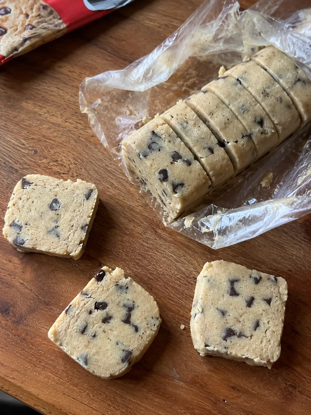 A roll of refrigerated chocolate chip cookie dough scored into slices, with three slices cut onto a wooden board