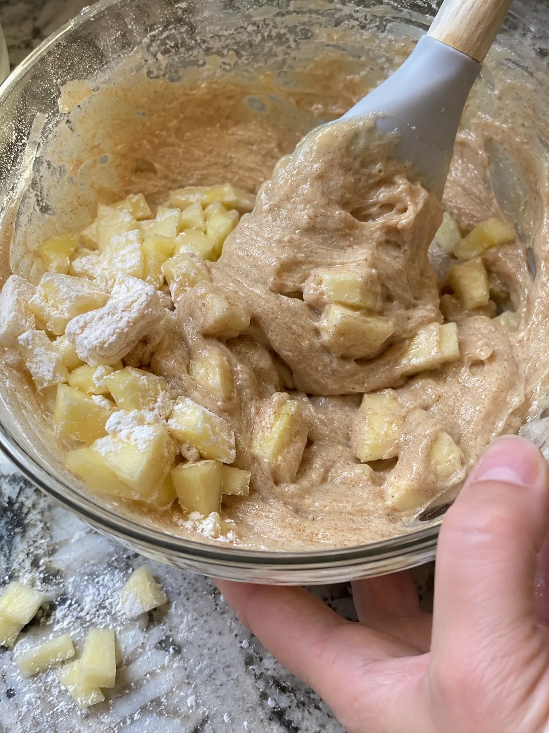 Flour-dusted apple cubes being folded into a spiced cake batter with a spatula