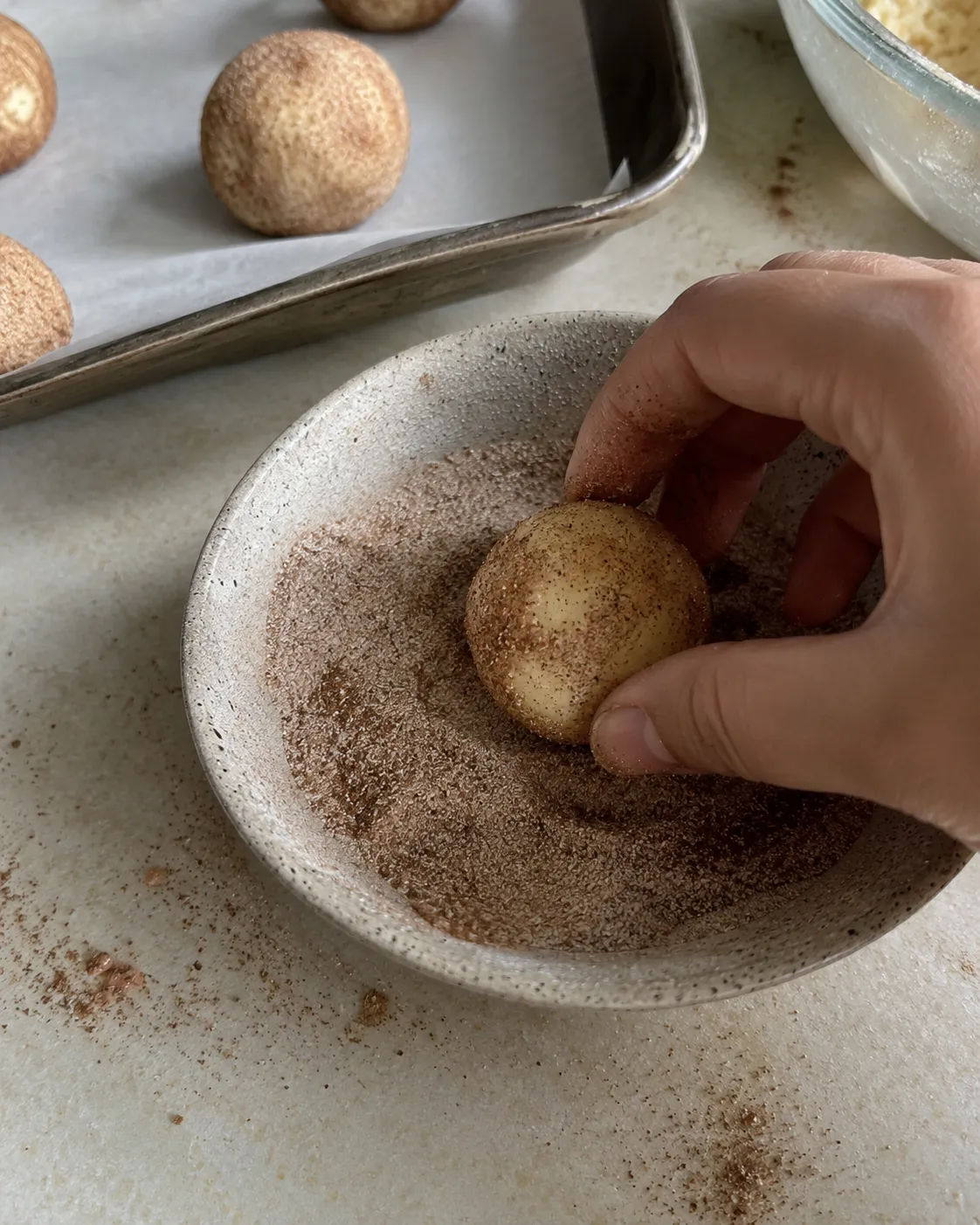 A hand rolling a ball of snickerdoodle dough in a bowl of cinnamon sugar