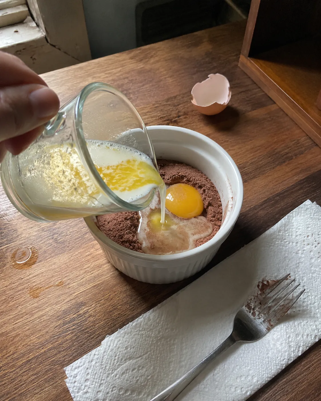 Melted butter and milk being poured onto the dry cocoa mixture in a ramekin, with an egg yolk sitting on top