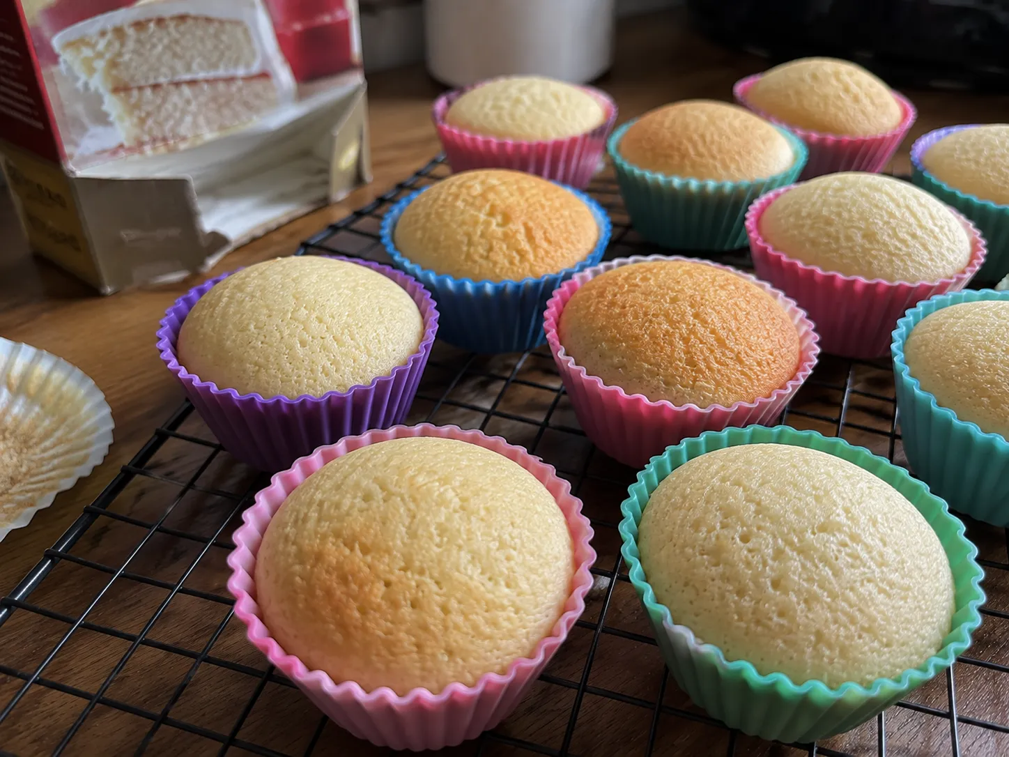 Cupcakes baked from a box mix in colored silicone cups, cooling on a wire rack beside the empty box