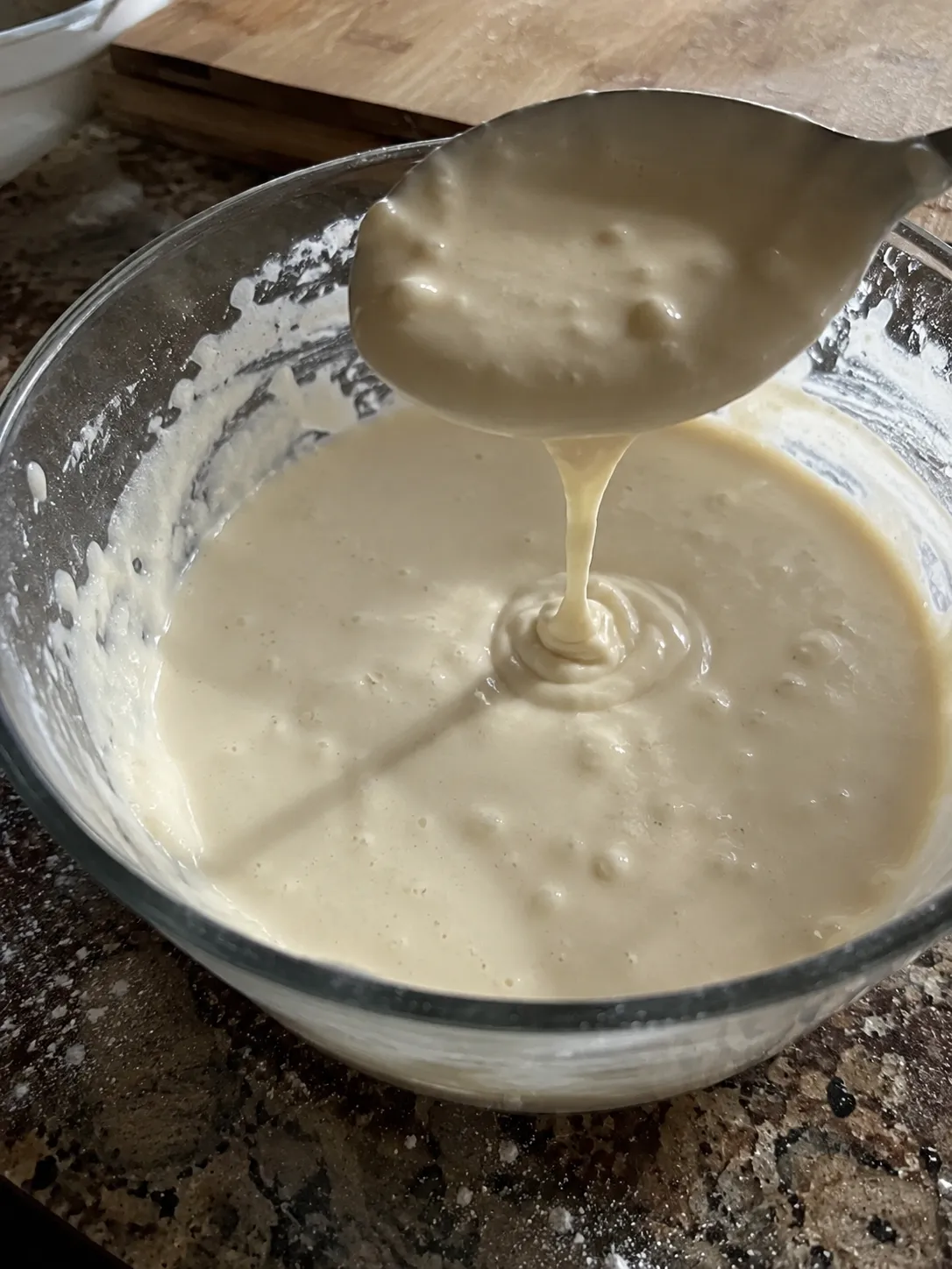 Funnel cake batter falling from a ladle in a thick ribbon into a glass mixing bowl