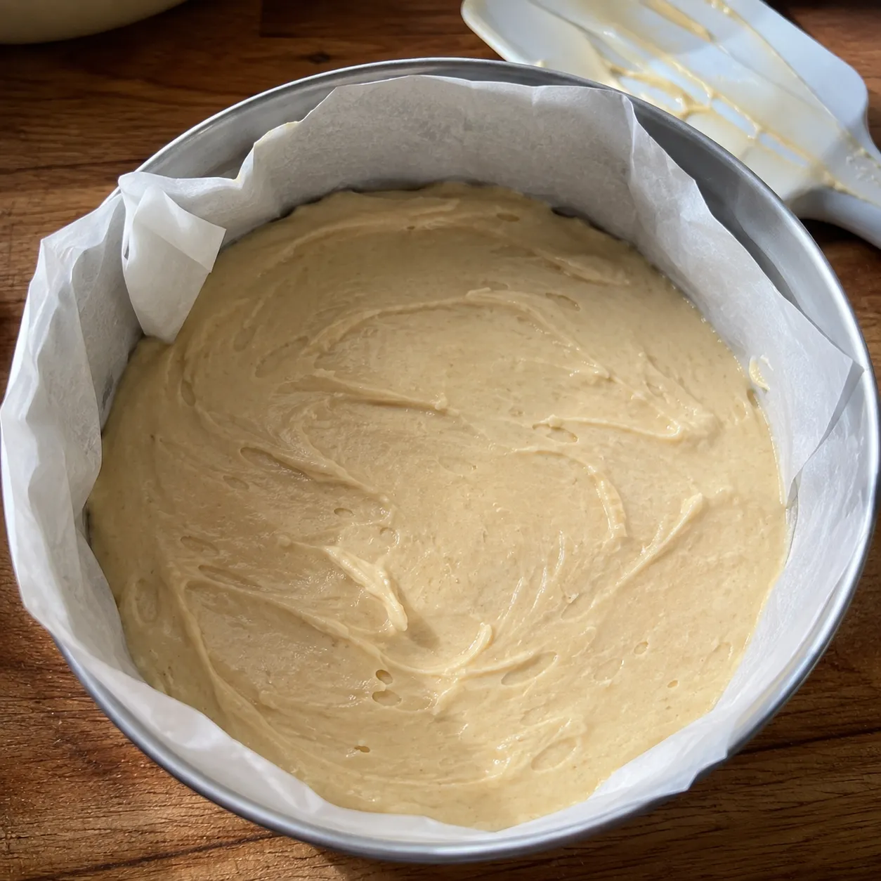 Pale cake batter levelled in a parchment lined round pan with a spatula resting alongside