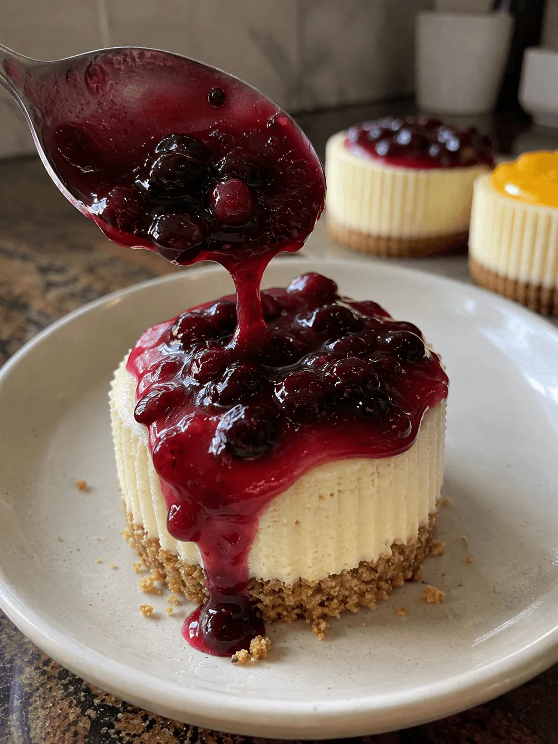 Blueberry compote being spooned over a mini cheesecake on a plate with a lemon curd topped one behind