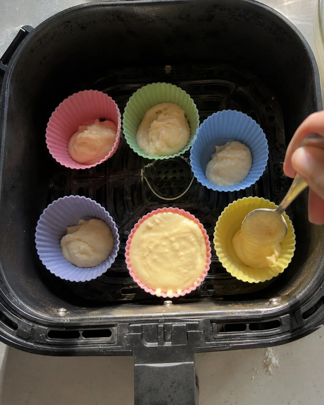 Six pastel silicone cupcake cups arranged around an air fryer basket with vanilla batter being spooned into them