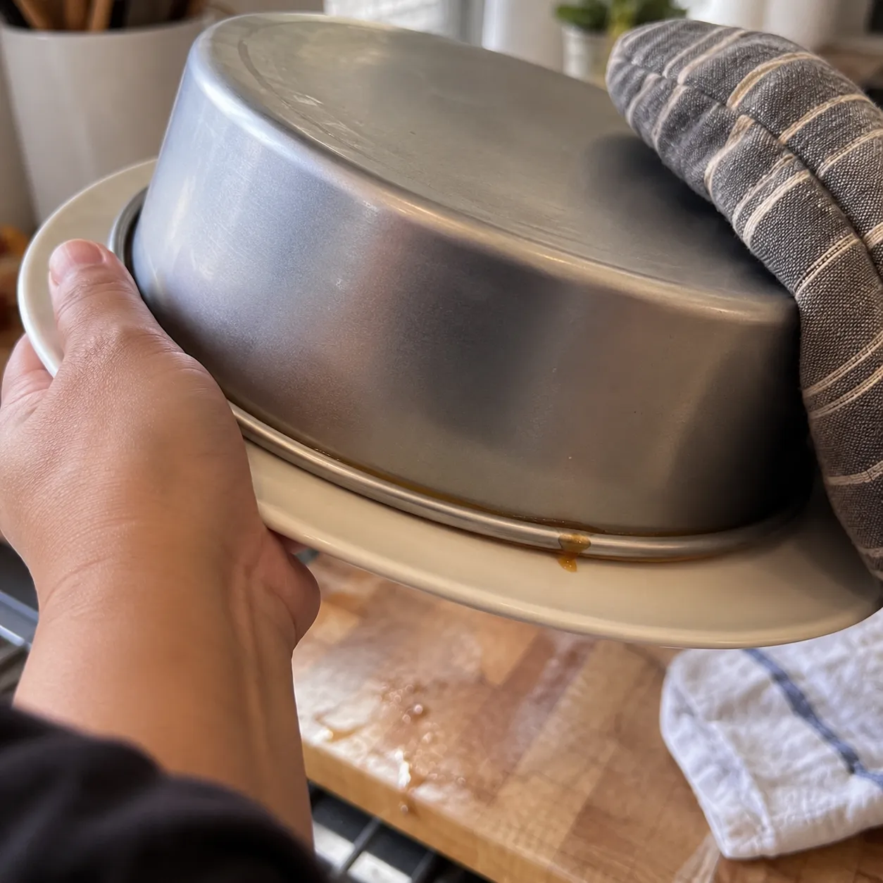 Two hands turning a cake pan upside down onto a serving plate, one hand steadying the plate and the other holding the hot pan through a folded towel, syrup running onto the plate