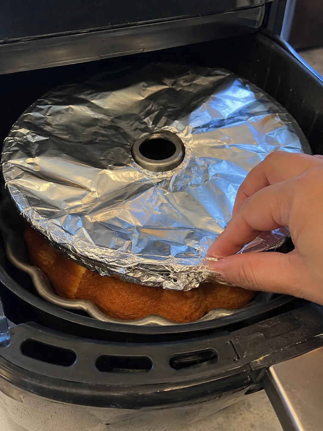 A hand pressing a disc of foil down over the rim of a bundt pan inside an air fryer basket, the risen cake visible underneath