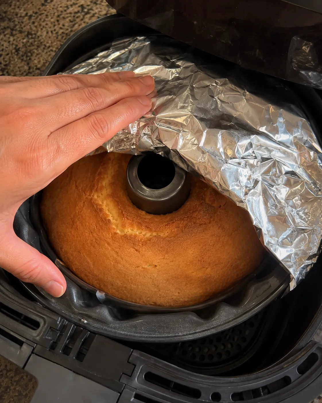 A hand laying a sheet of foil over a browned pound cake in a tube pan sitting inside an air fryer basket