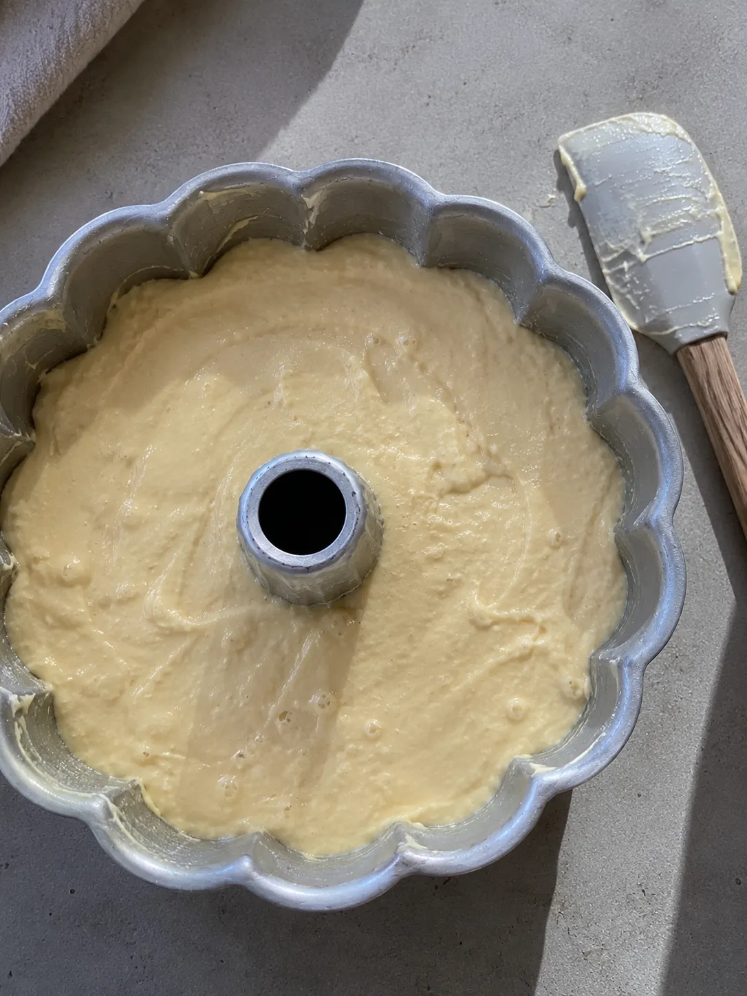 Pale cake batter smoothed into a greased fluted bundt pan, filled to about two thirds, with a spatula alongside