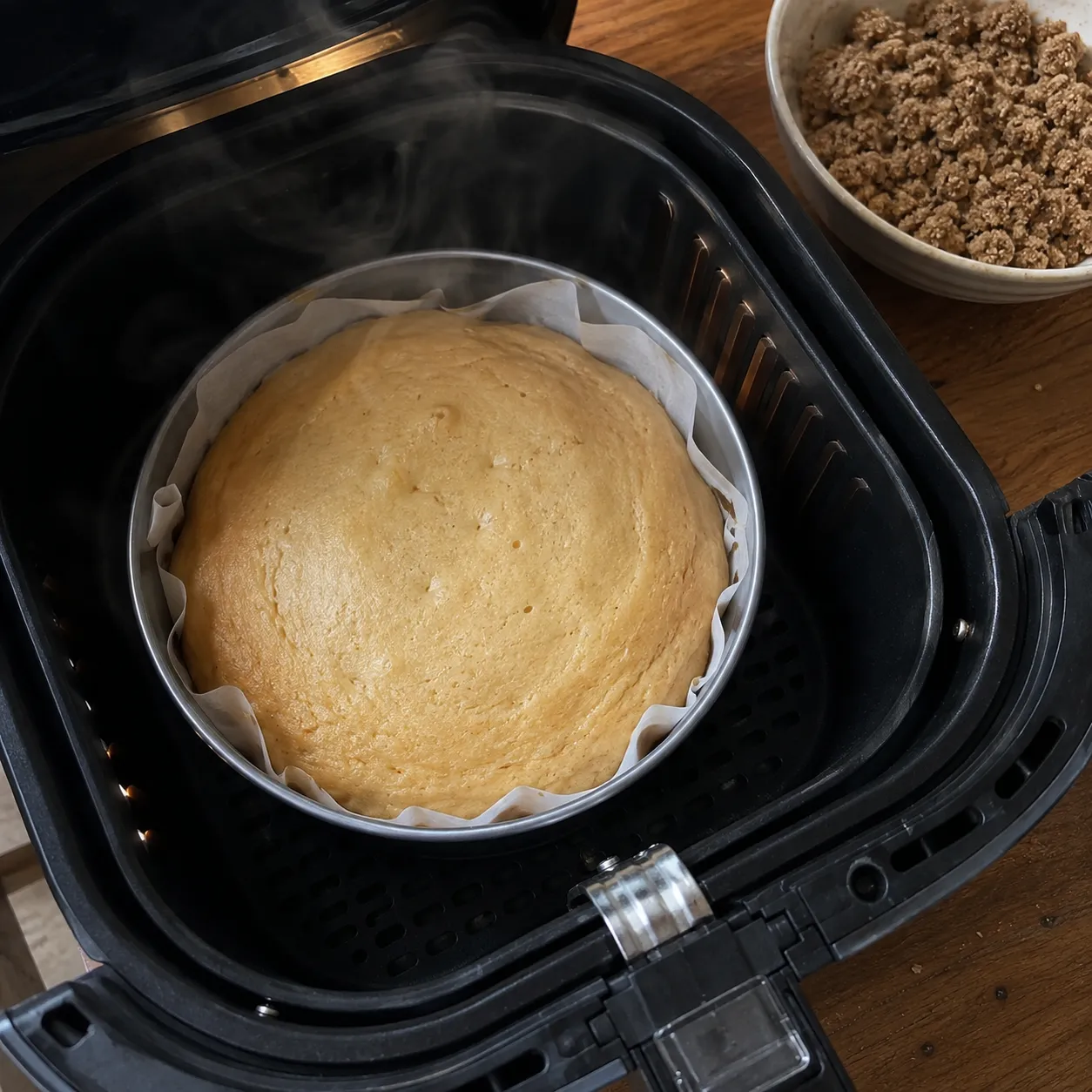 A risen pale cake in a parchment lined pan inside an open air fryer basket, with a bowl of streusel waiting on the counter beside it