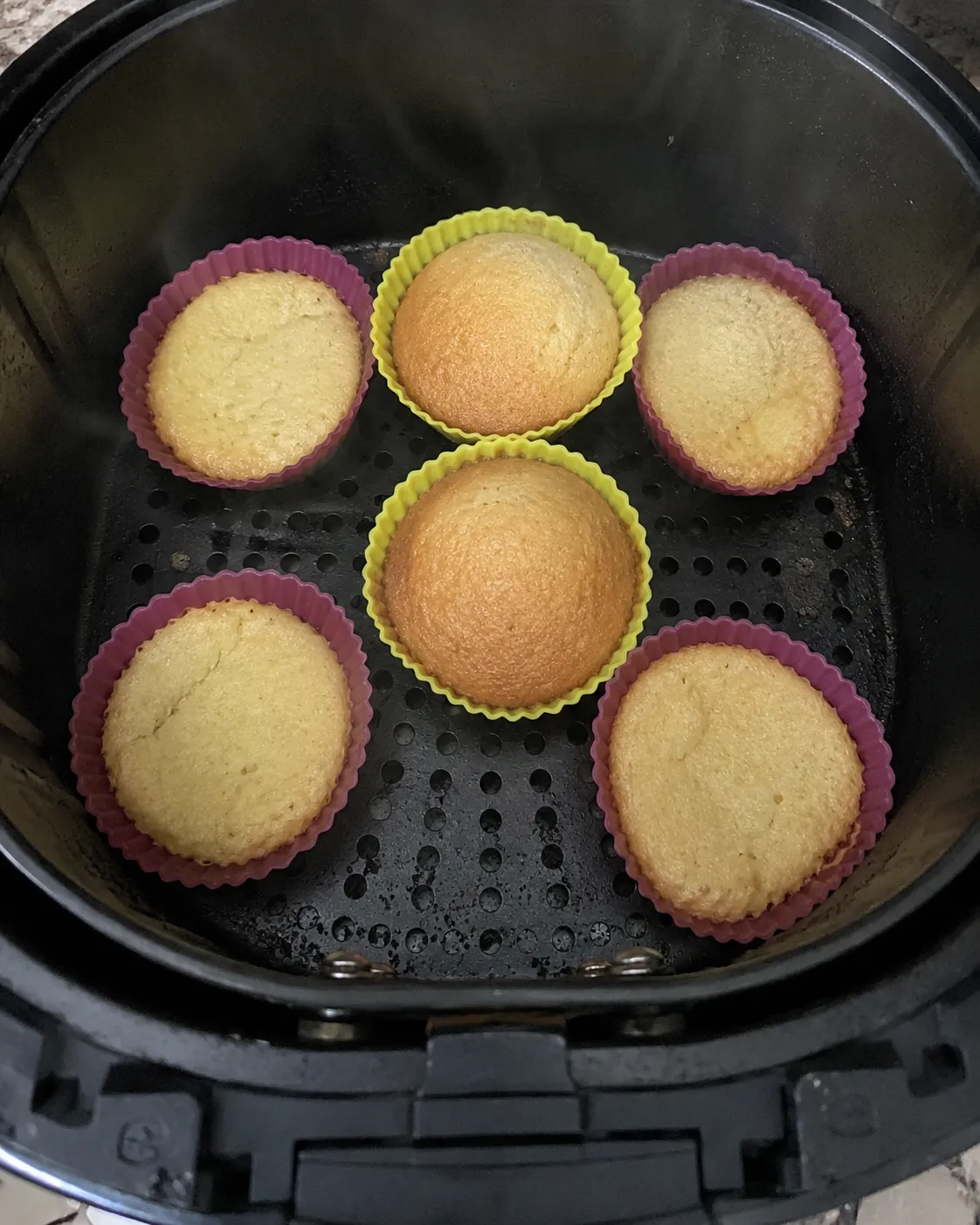 Six baked cupcakes in silicone cups in a round air fryer basket, the two down the middle domed higher and browner than the four around the edge