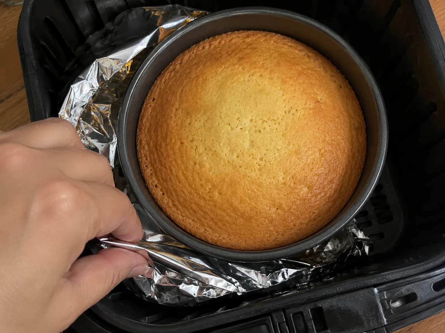 A baked round cake in its pan sitting in an air fryer basket on a foil sling