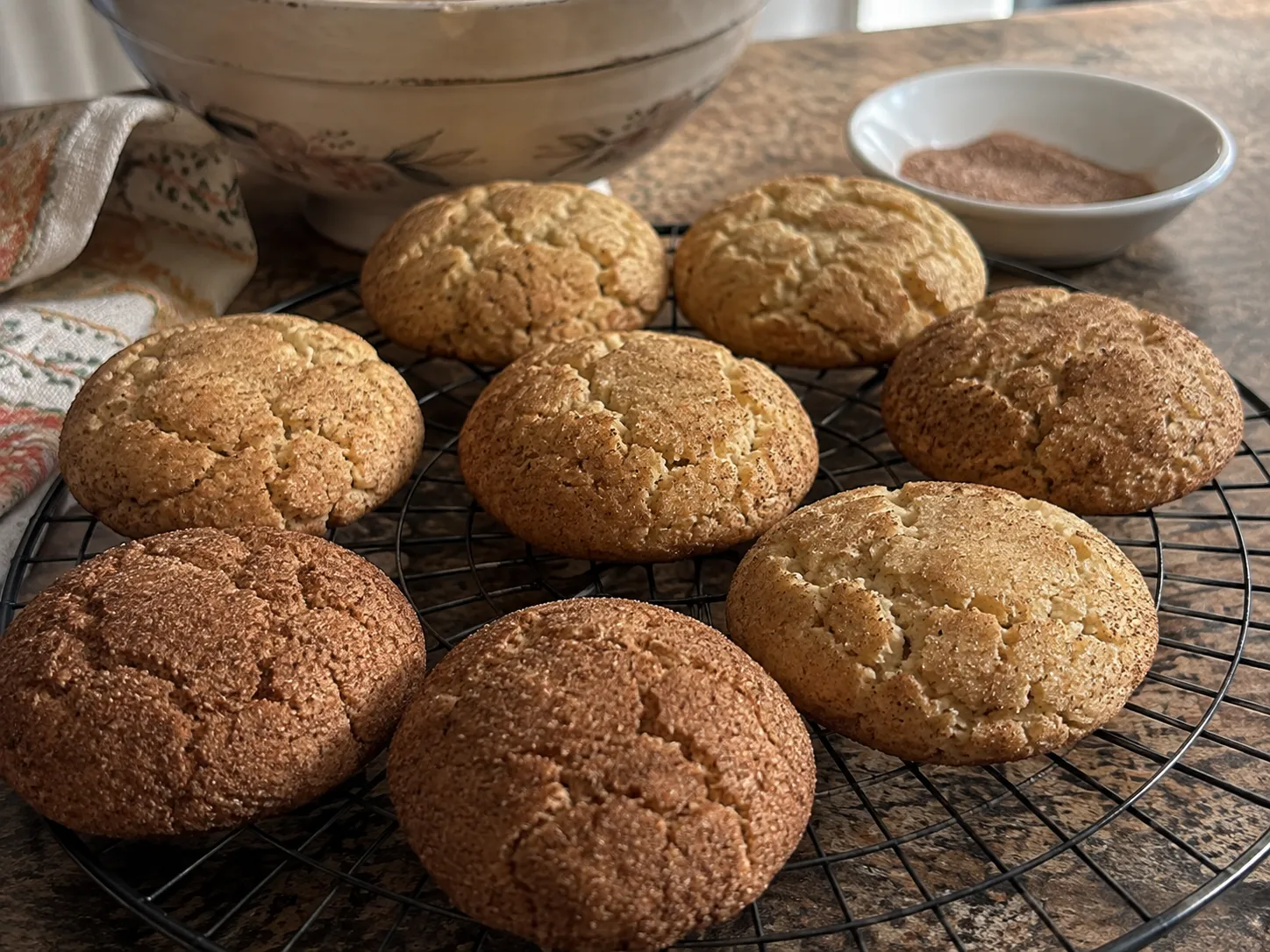 Eight baked snickerdoodles cooling on a wire rack beside a bowl of cinnamon sugar