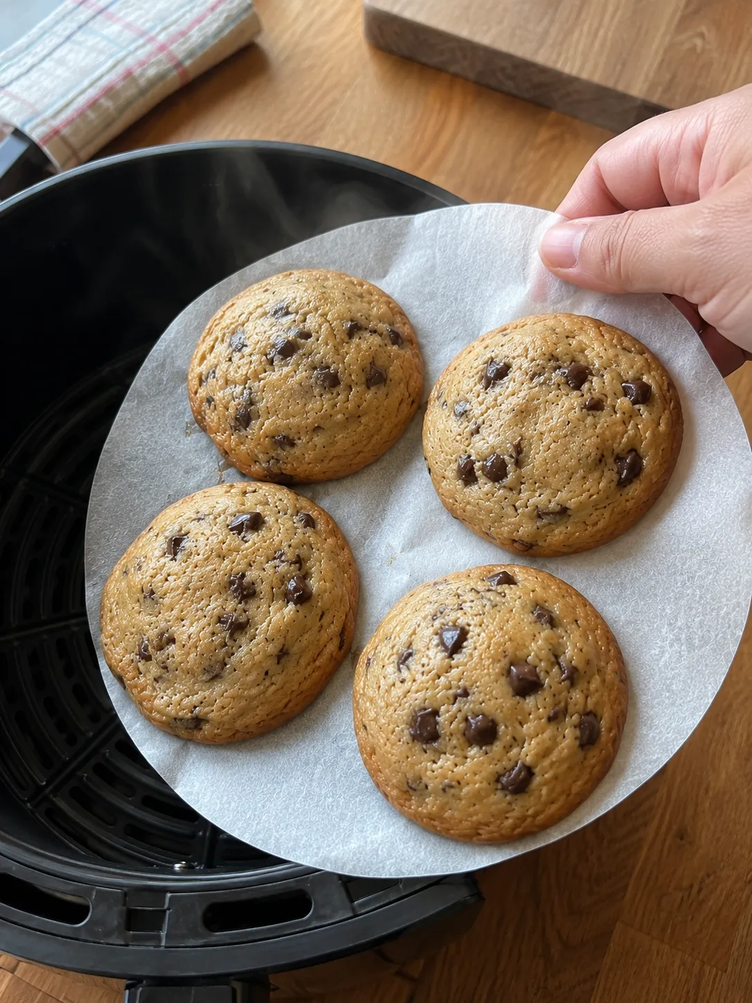 Lifting four finished cookies out of an air fryer basket on the parchment round