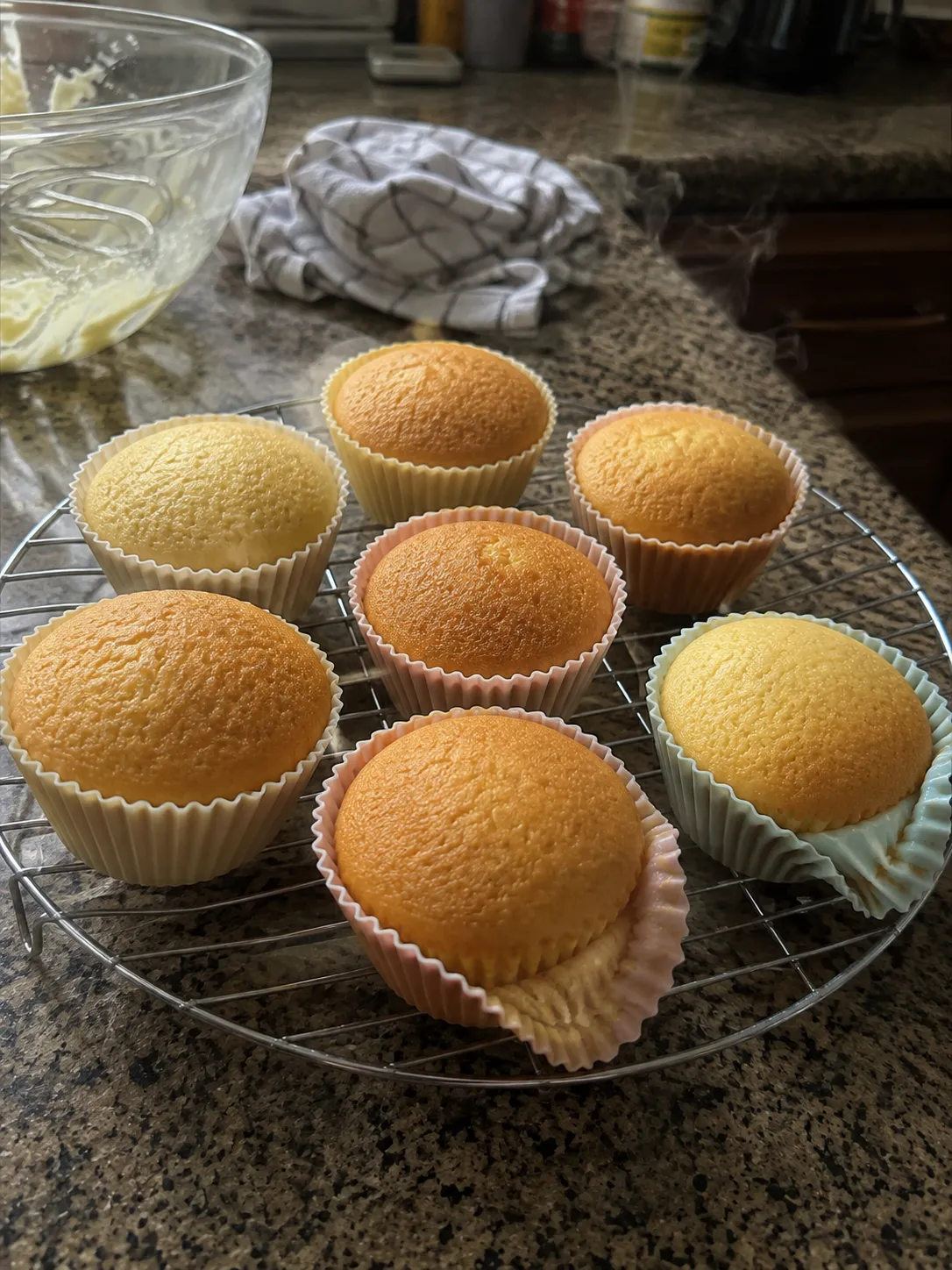 Seven unfrosted cupcakes in pastel silicone cups steaming on a wire cooling rack on a granite counter
