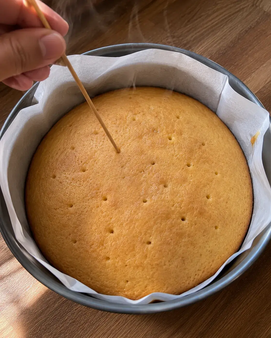 A skewer poking a hole into a freshly baked lemon cake still in its pan, with steam rising