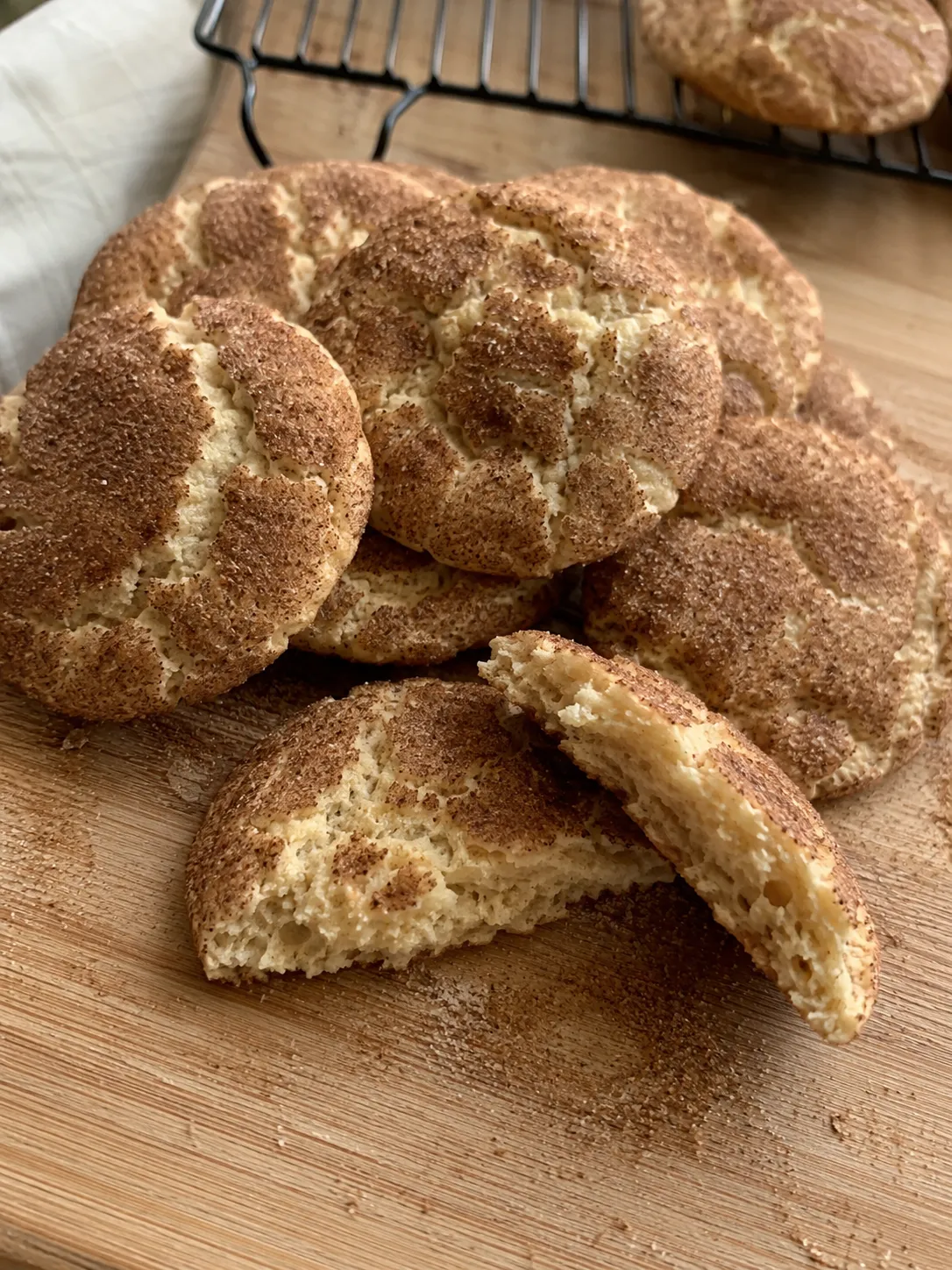 Air fryer snickerdoodles piled on a wooden board with one broken open to show the soft crumb