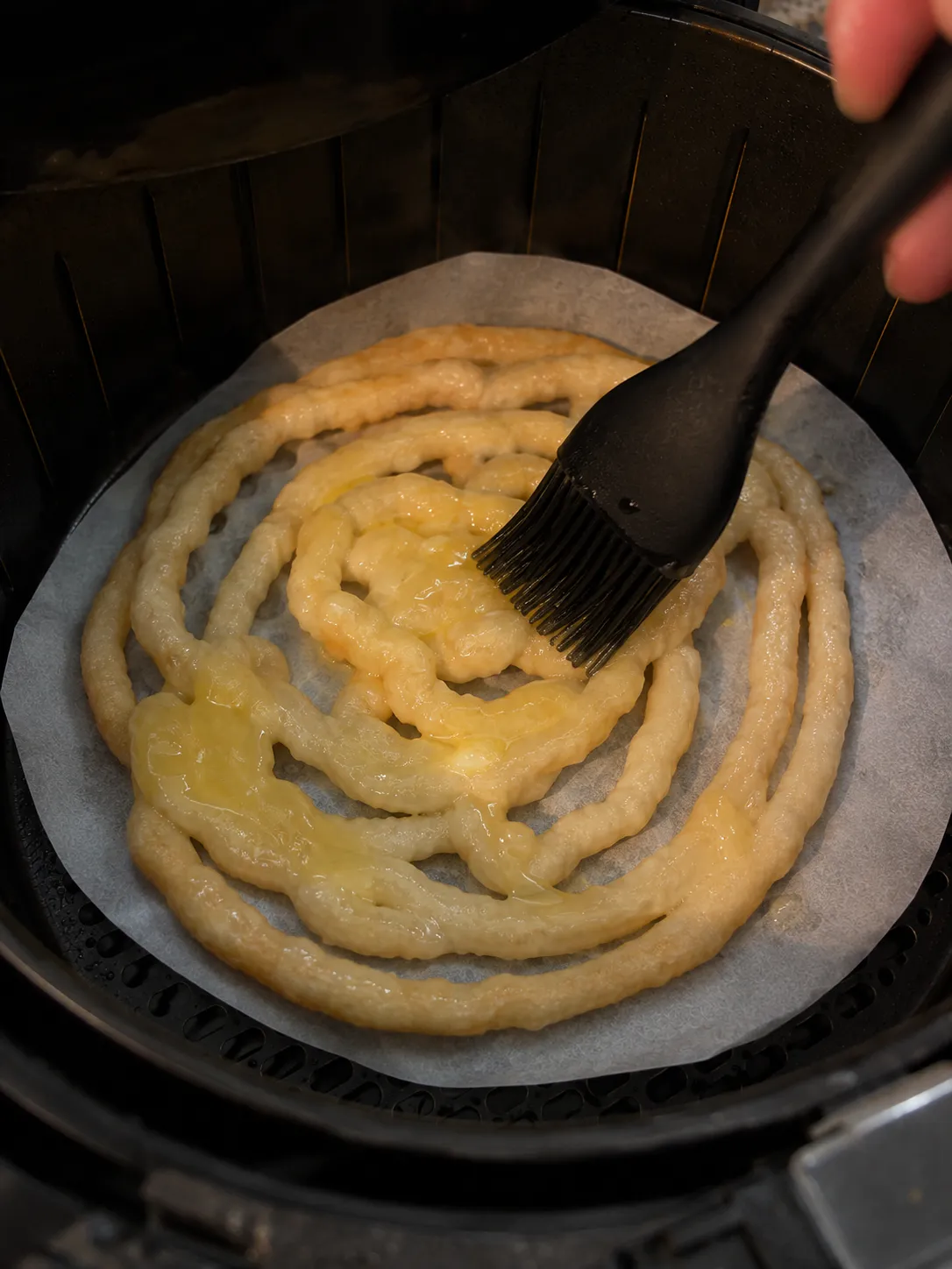 Melted butter being brushed onto a piped funnel cake in an air fryer basket with a silicone brush