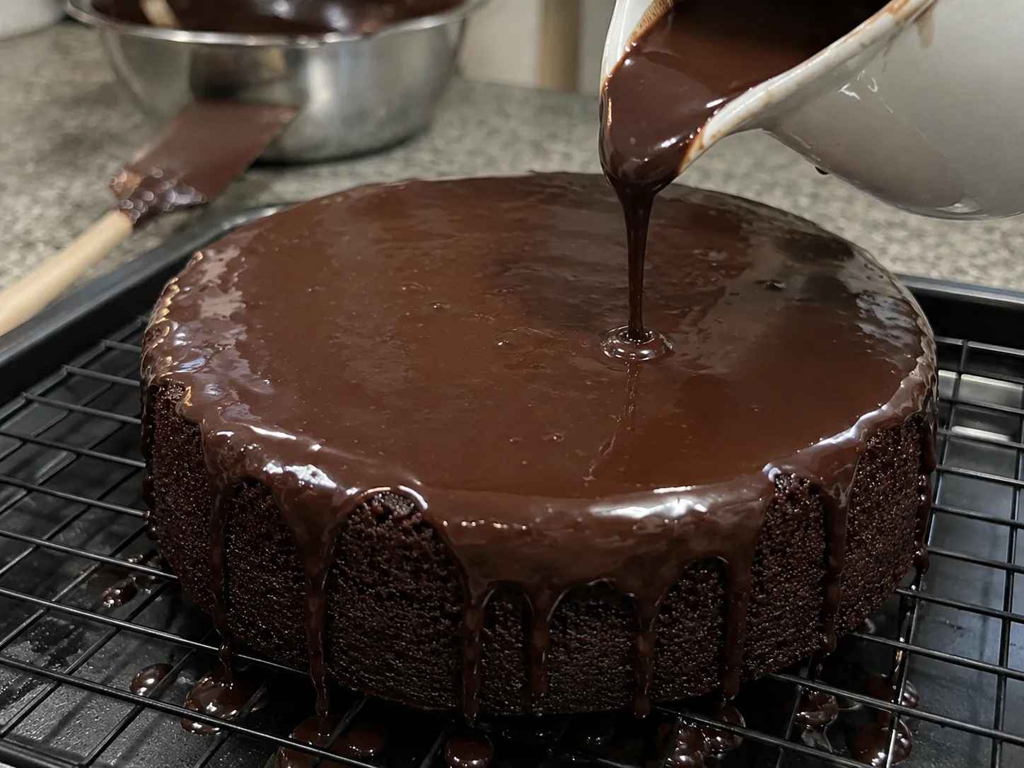 Dark chocolate ganache being poured from a jug over a cooled chocolate cake sitting on a wire rack above a baking tray