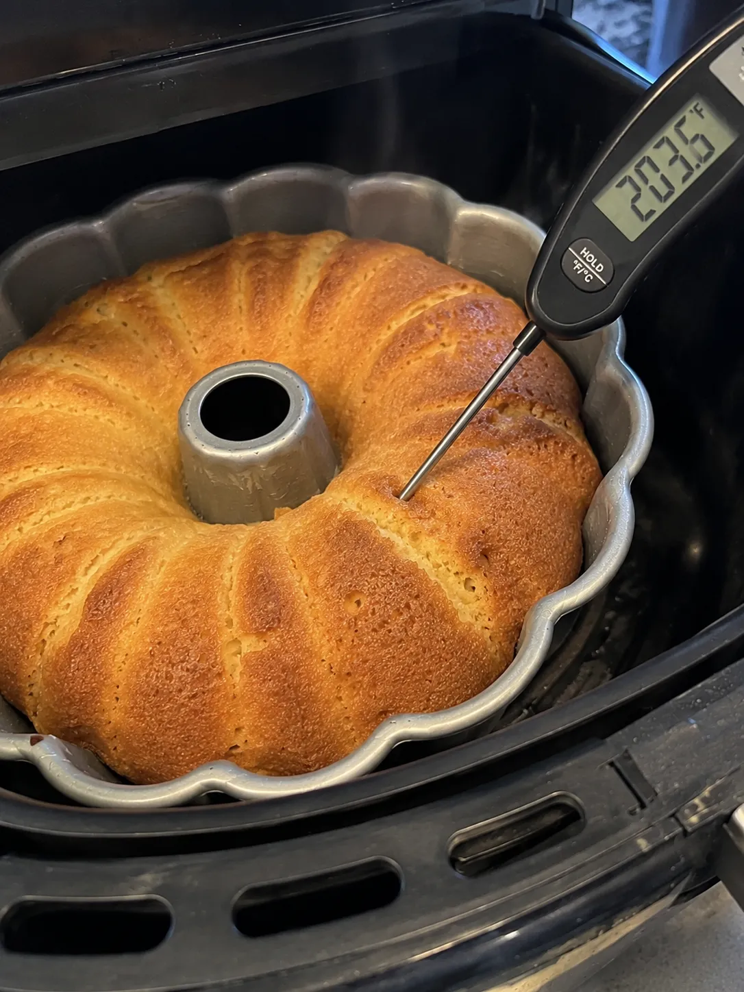 An instant-read thermometer angled into the thick part of a golden bundt cake in an air fryer basket, the display reading 203.6 degrees Fahrenheit