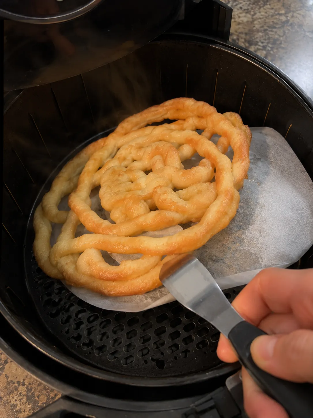 A set funnel cake being lifted off its parchment round with a thin spatula inside an air fryer basket