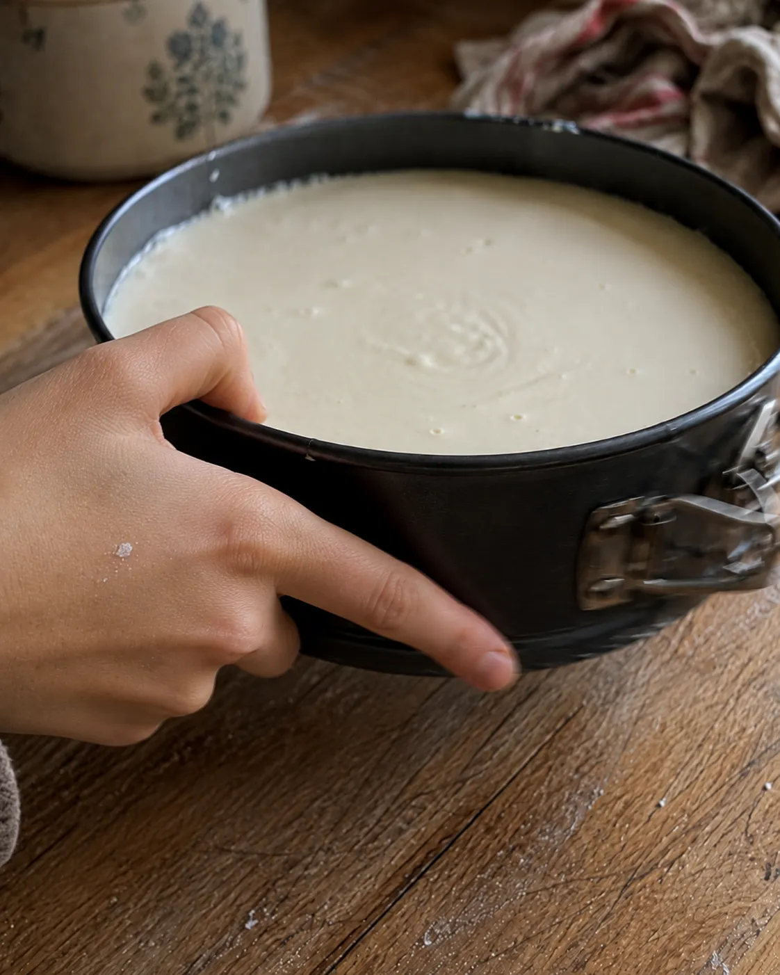 Two hands holding a springform pan filled with smooth cheesecake batter