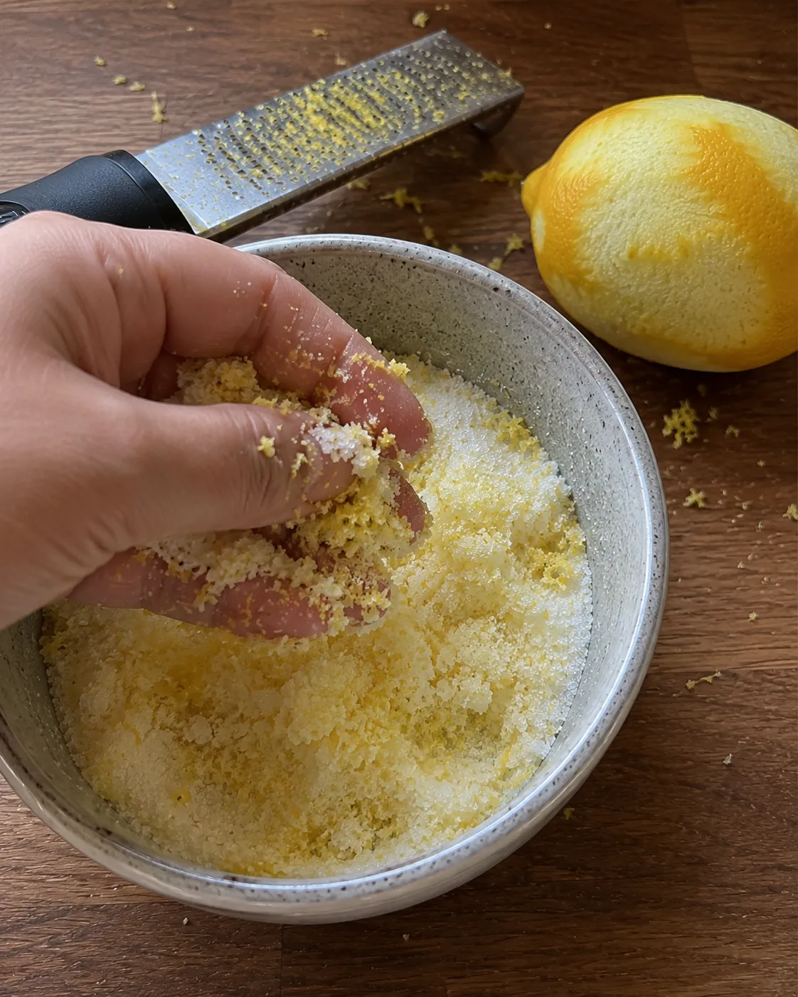 A hand rubbing lemon zest into granulated sugar in a bowl, the sugar turned damp and pale yellow