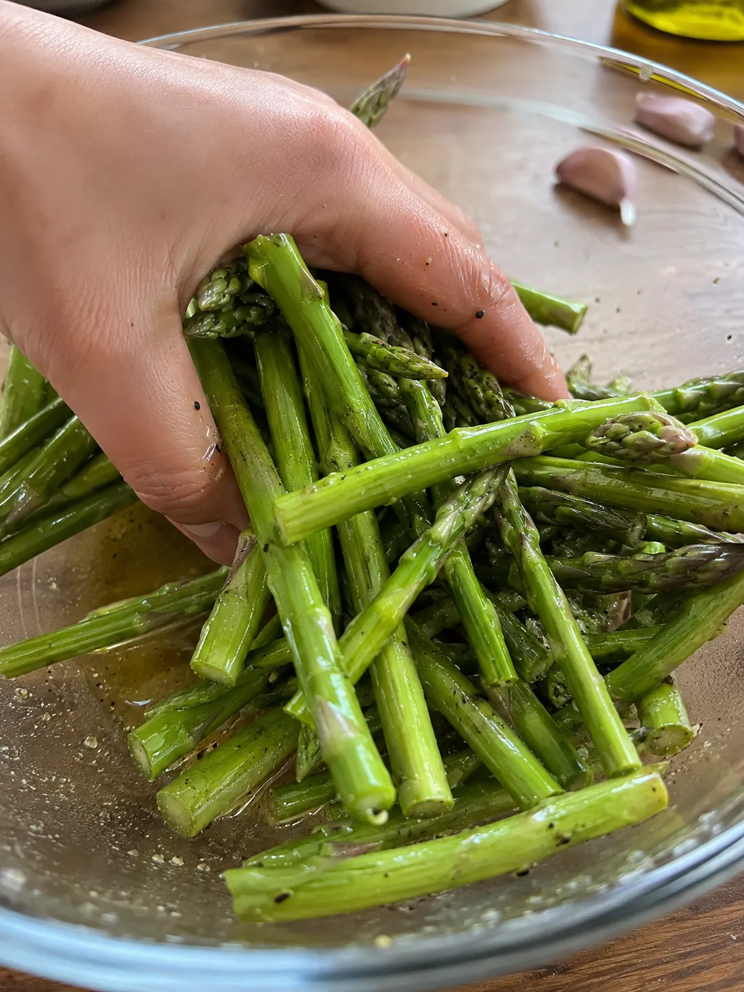 A hand tossing trimmed asparagus with oil and black pepper in a glass bowl