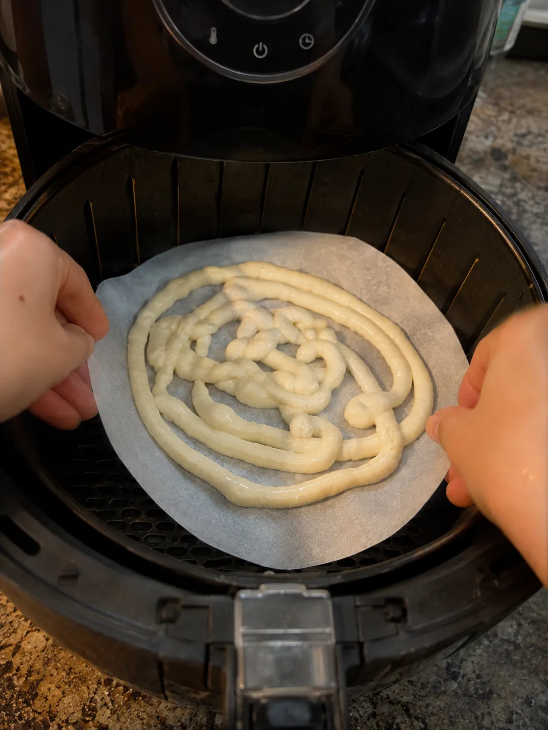Two hands lowering a parchment round of piped funnel cake batter into an air fryer basket