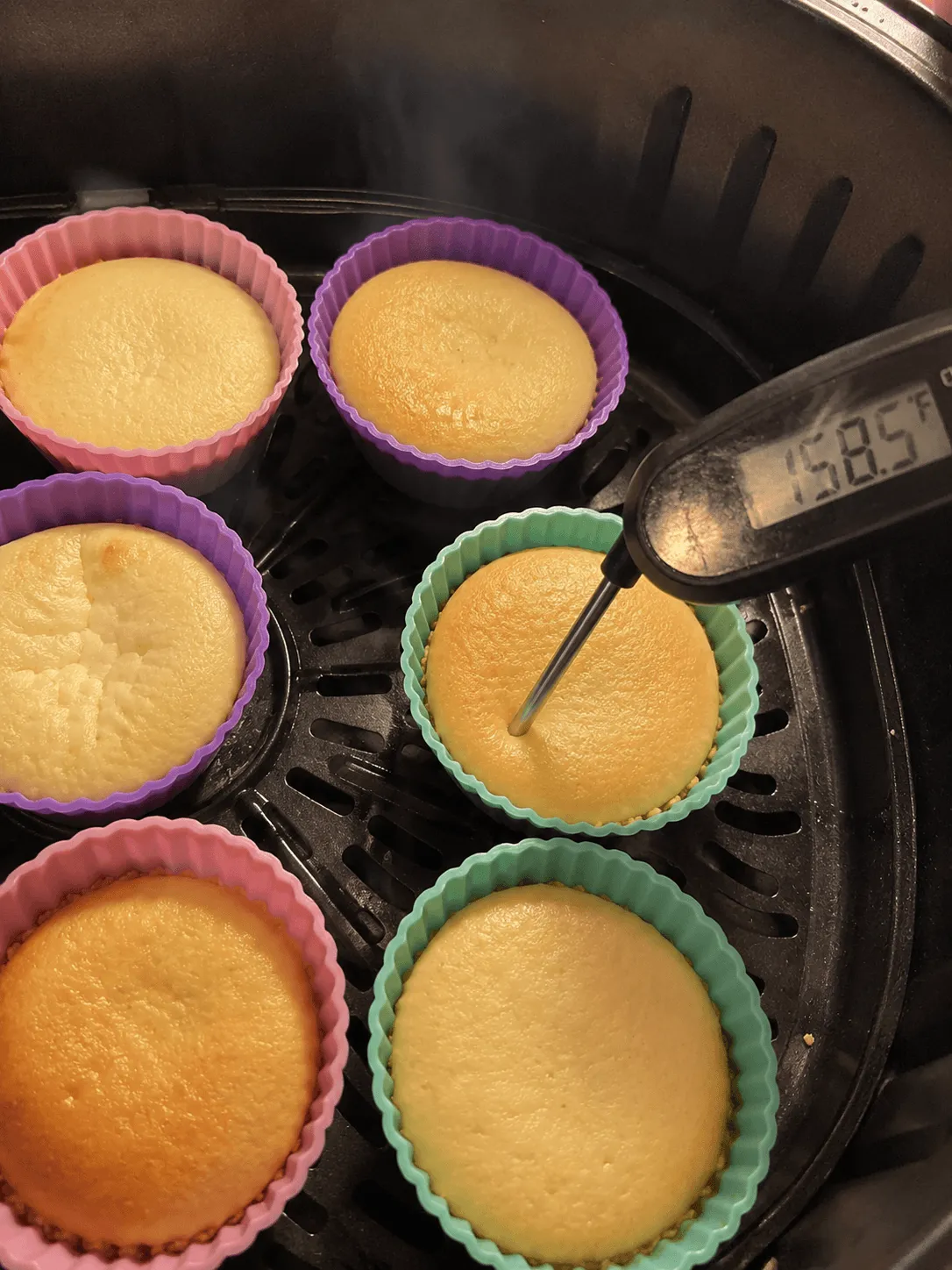 Six mini cheesecakes in silicone cups in an air fryer basket with a thermometer reading 158.5 degrees Fahrenheit