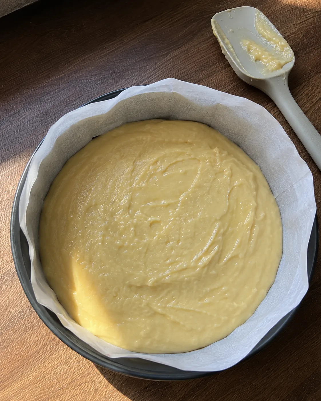 Lemon cake batter scraped into a parchment-lined 6-inch round pan and leveled, with a spatula resting nearby