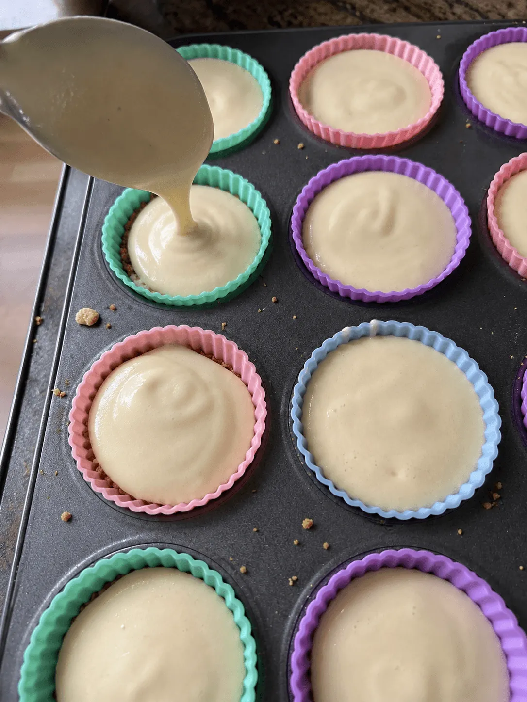 Cheesecake batter being poured from a jug into crust-lined silicone cups in a muffin tin