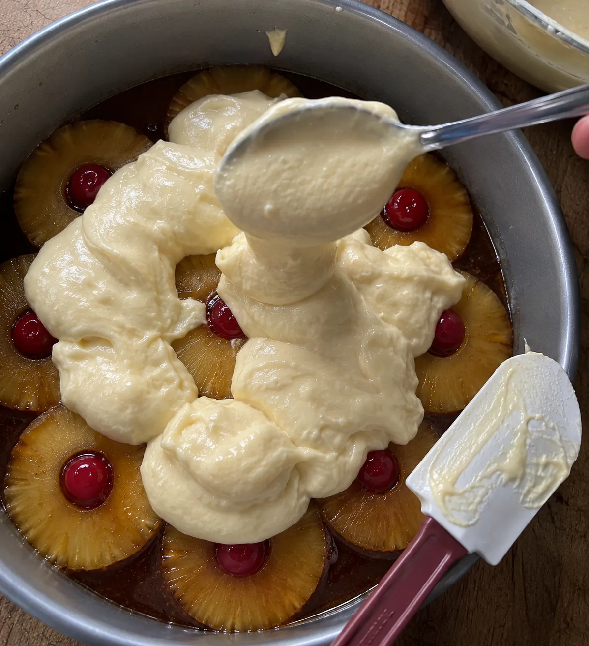 A spoon dolloping pale cake batter over pineapple rings and cherries set in dark caramel in a cake pan