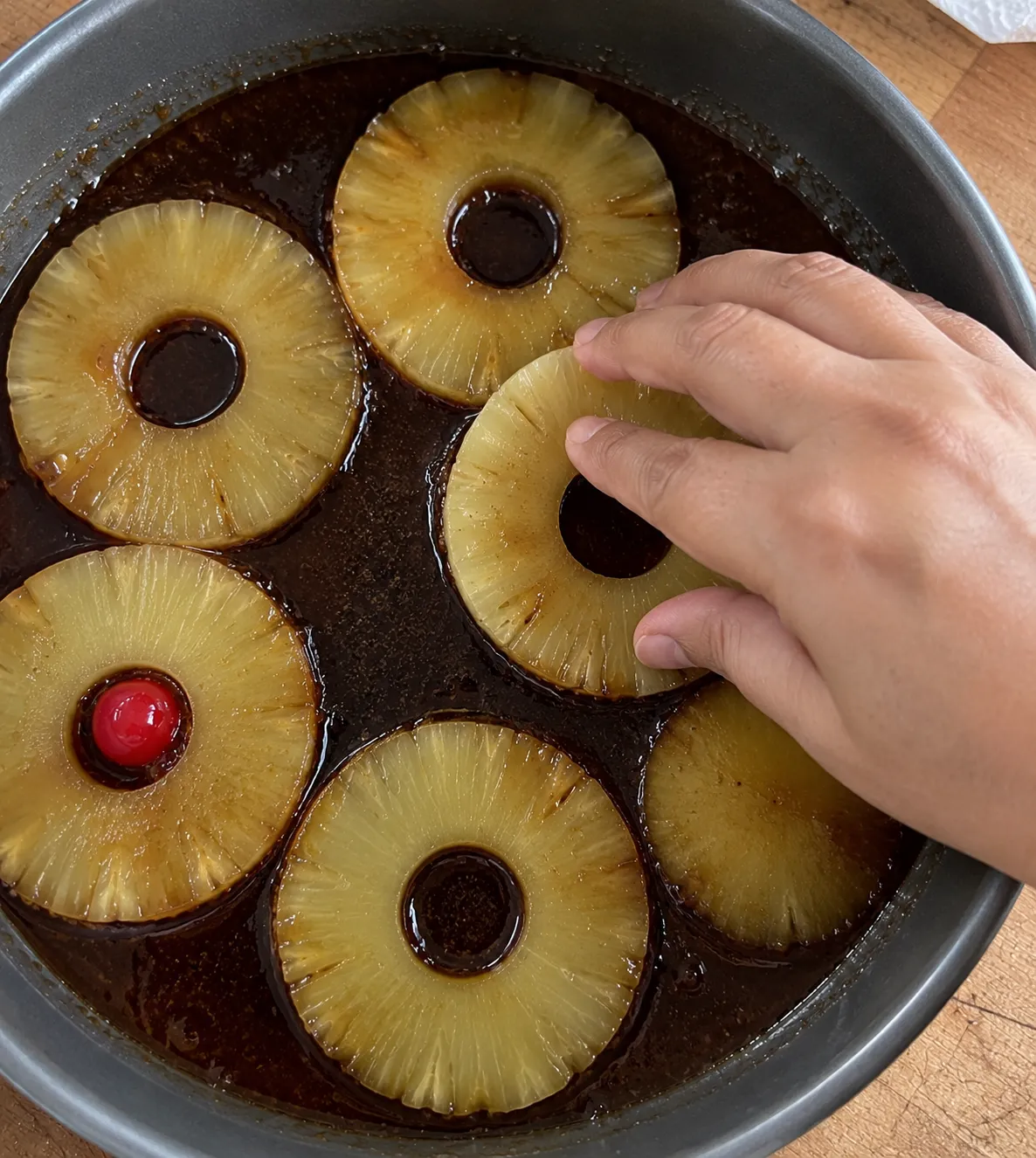 A hand arranging blotted pineapple rings into dark bubbling caramel in an ungreased-looking metal cake pan, one cherry already placed