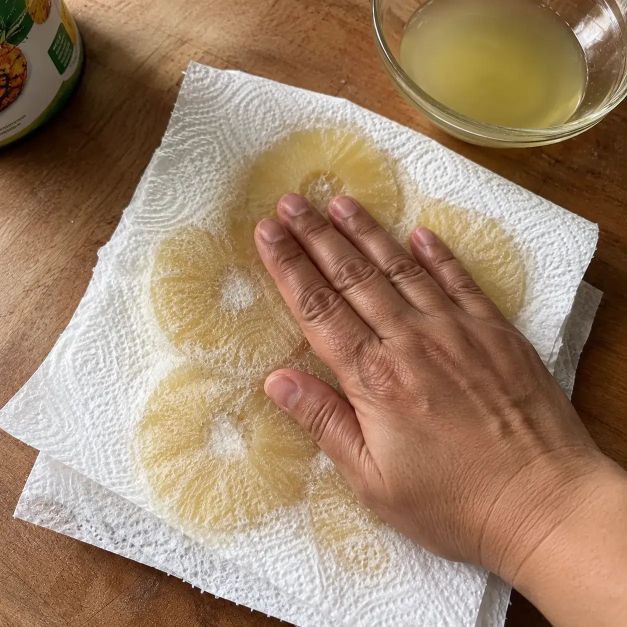 A hand pressing a paper towel down over canned pineapple rings, with wet patches visibly soaking through the towel