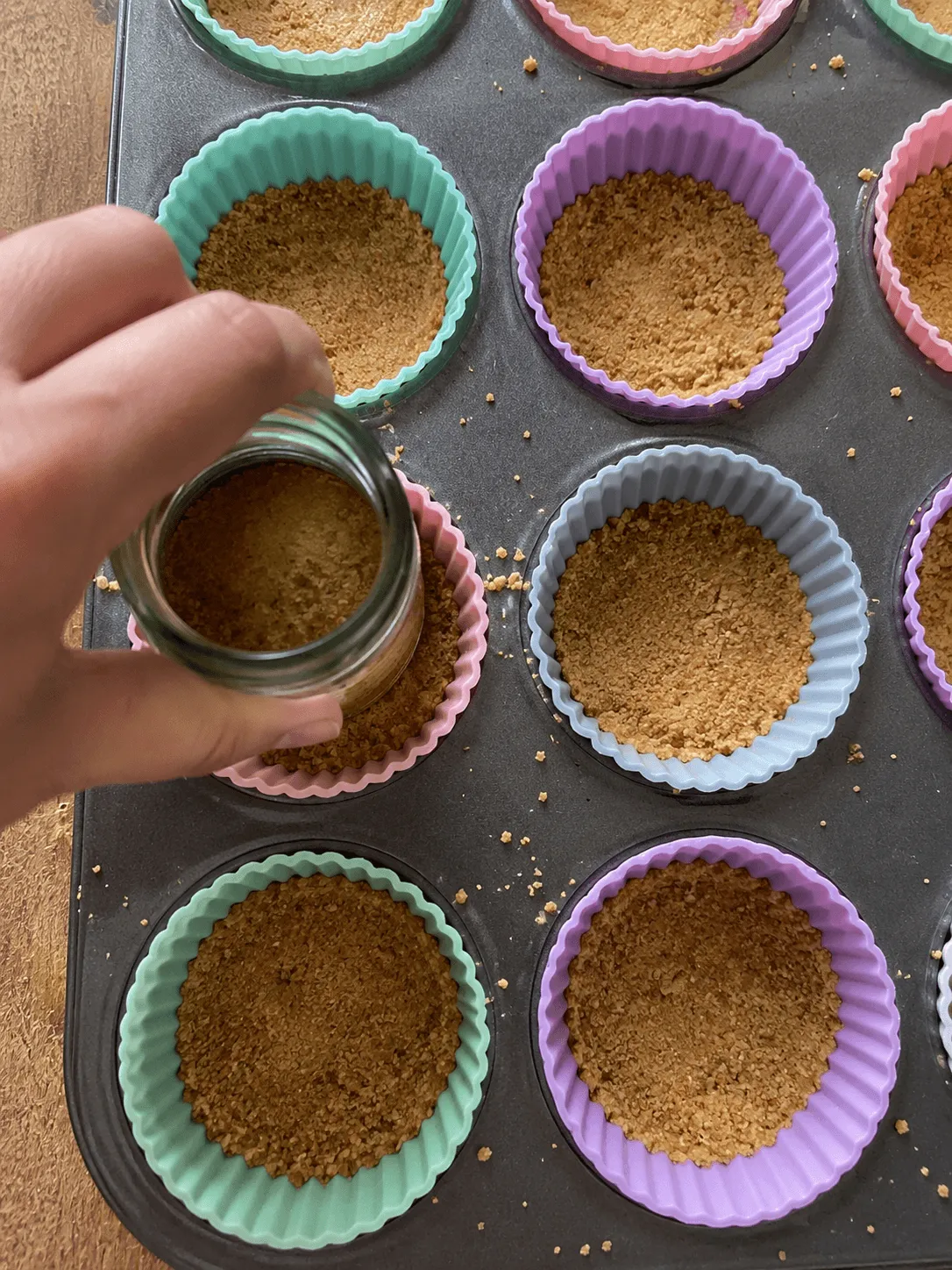 Graham crumb crust being pressed into pastel silicone cups standing in a metal muffin tin