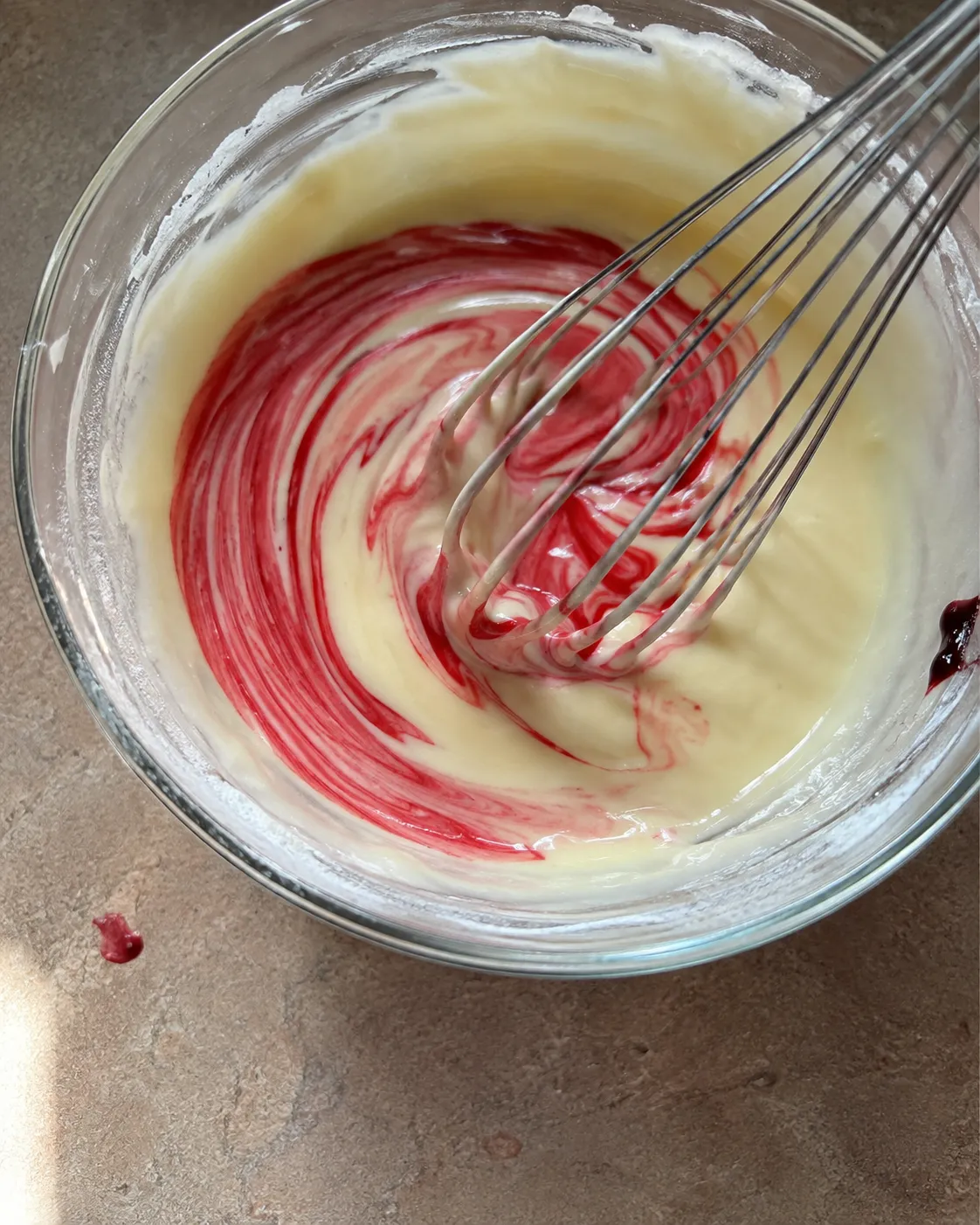Red gel food coloring being whisked into pale wet cake batter, still streaky and not yet fully combined