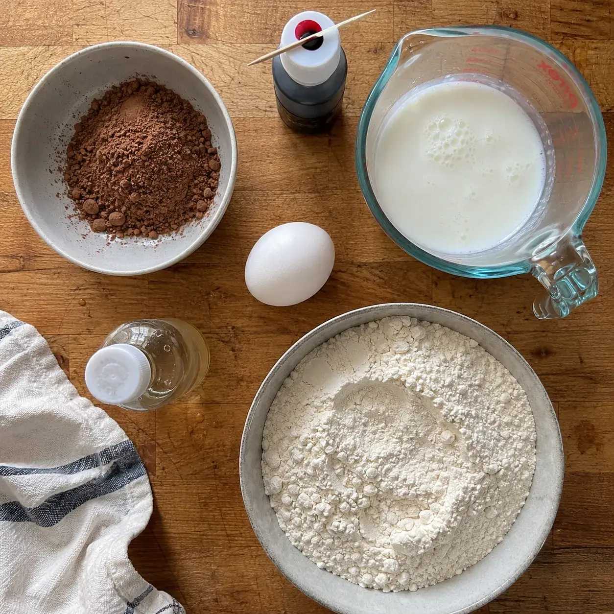 Ingredients for air fryer red velvet cake laid out on a wooden counter, including cocoa, buttermilk, egg, oil and flour